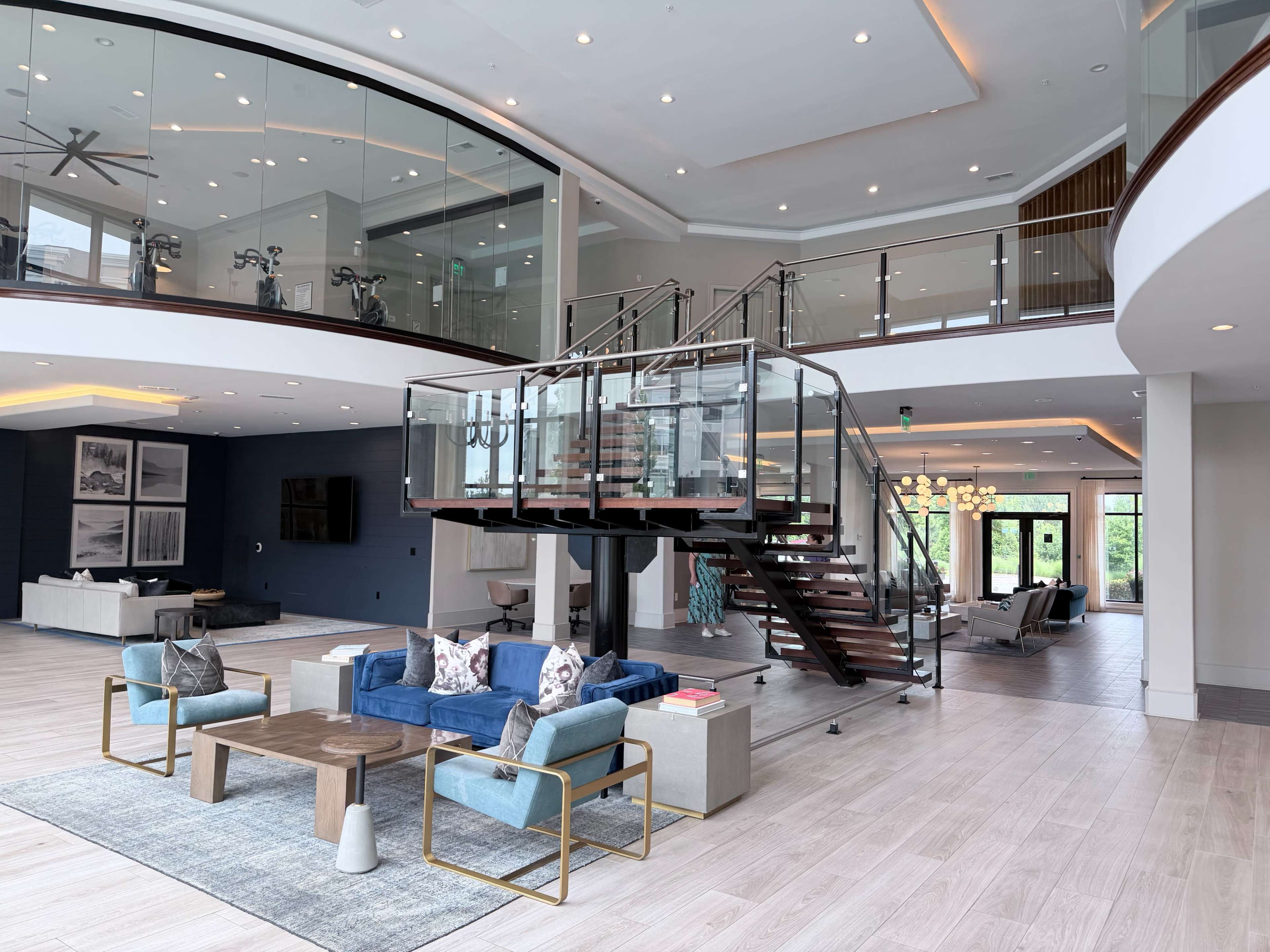 The image shows a spacious modern interior with an open floor plan, featuring a staircase with glass railings, a seating area with blue couches, and large windows allowing natural light.