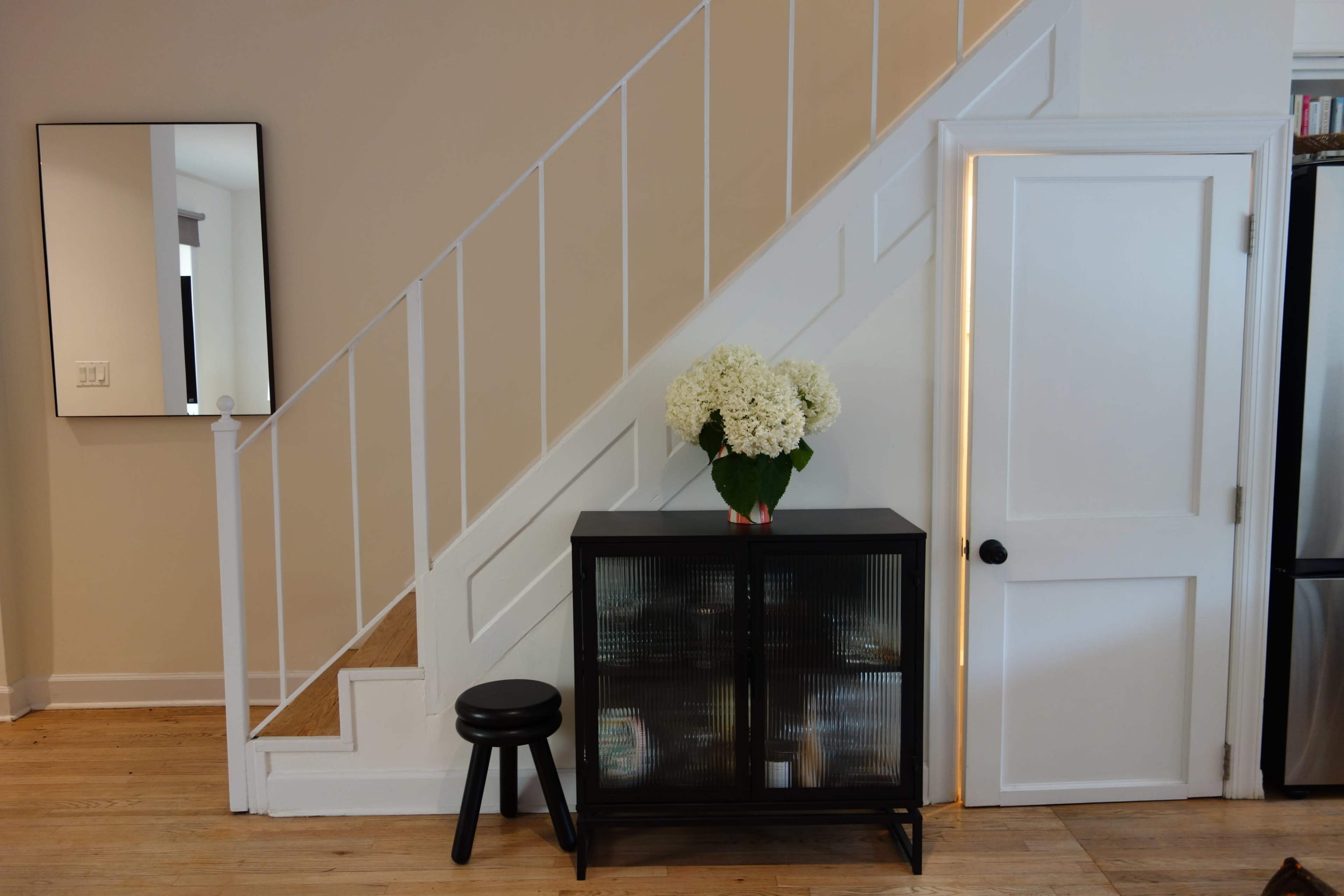 A black cabinet with a bouquet of white flowers sits next to a staircase, adjacent to a white door and a mirror on the wall.