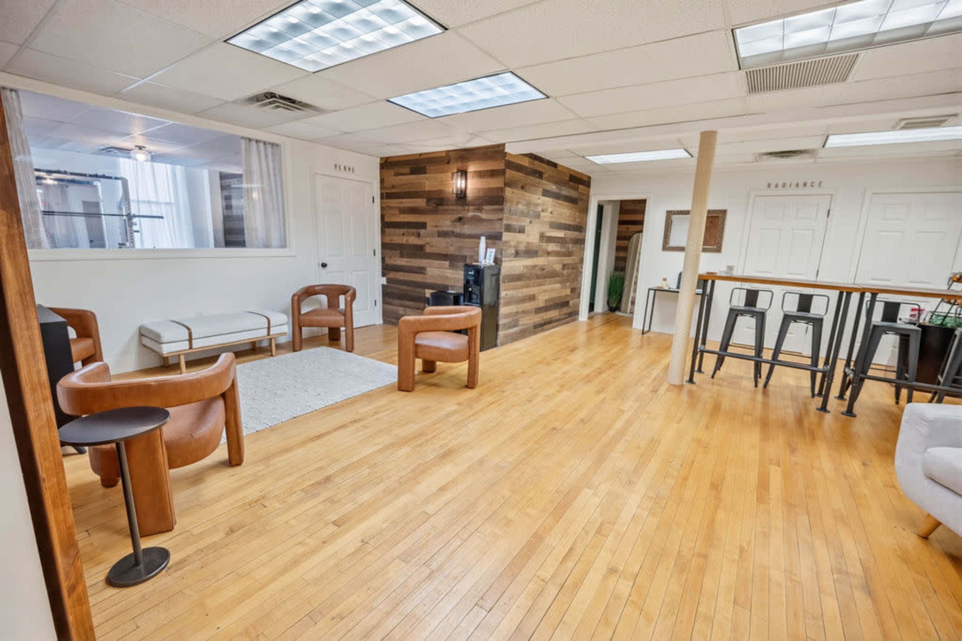 The image shows a spacious, well-lit interior with wooden flooring, featuring a seating area with modern chairs, a small rug, and a kitchenette.