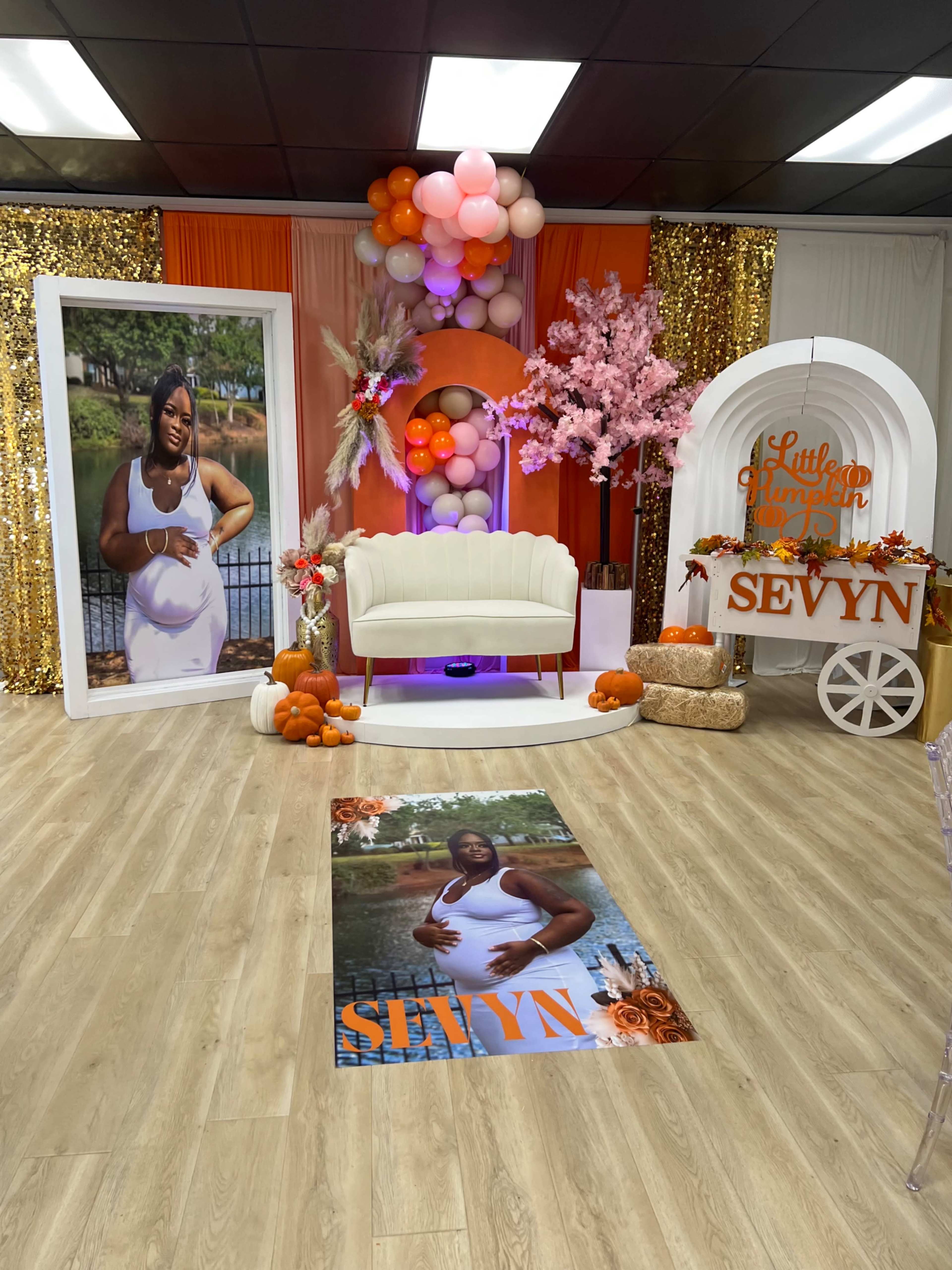 The image depicts a festive event space featuring a white couch, a large framed portrait of a woman, colorful balloons, and decorative elements like pumpkins and faux cherry blossom trees.
