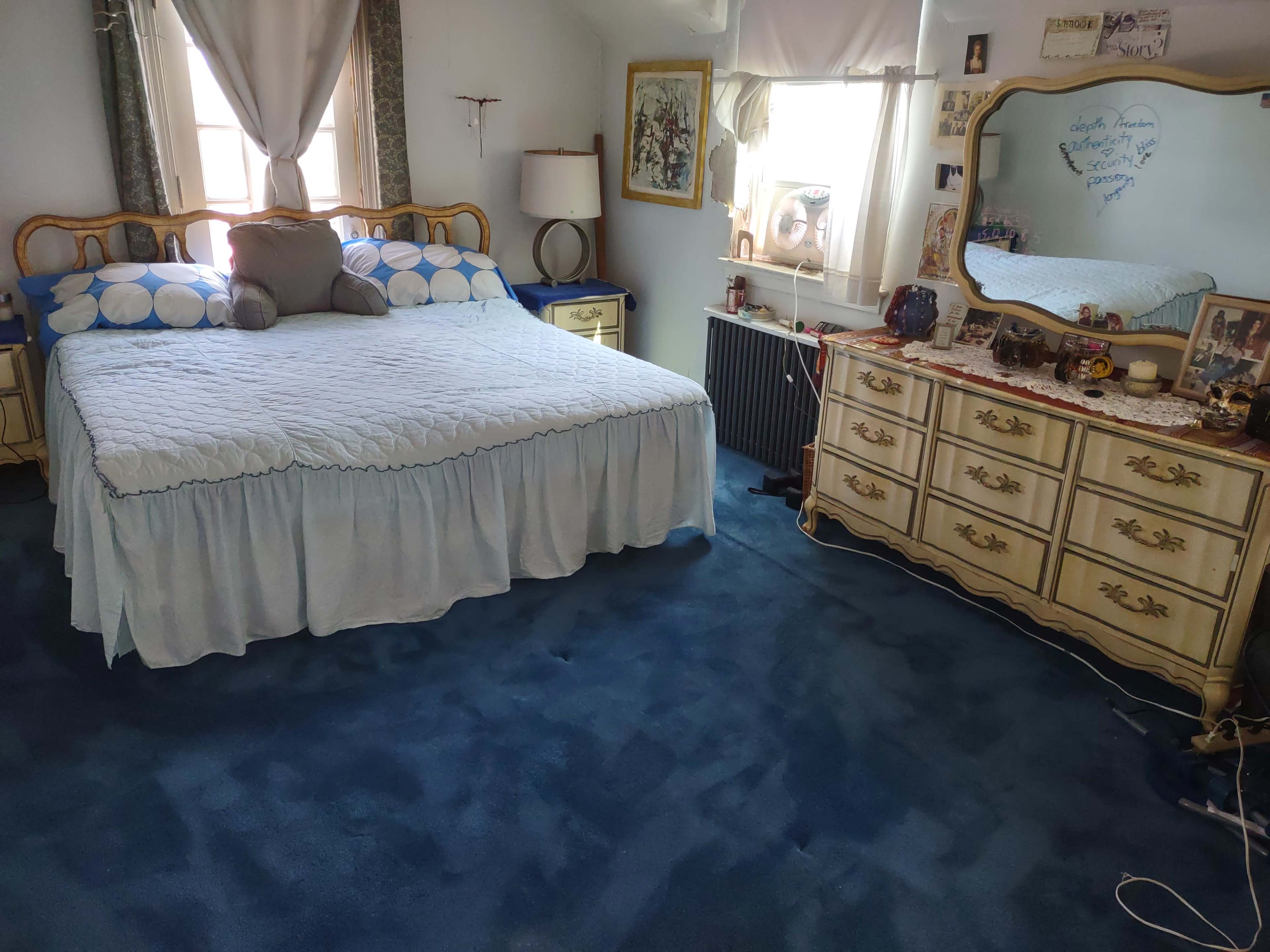 The room features a bed with a blue and white bedspread, a wooden dresser, and a large mirror, all situated on a blue carpet.