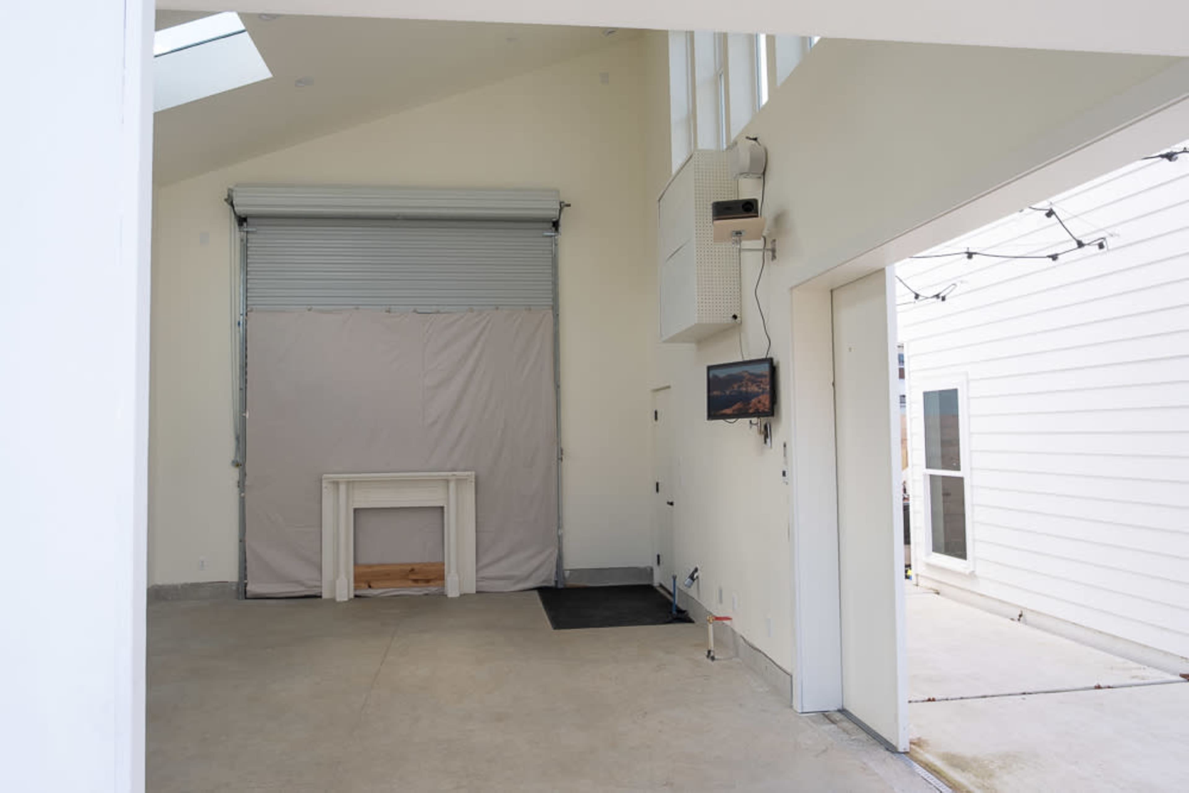 The image shows an empty interior space with a partially rolled-up garage door, a covered fireplace, and a mounted television on the wall.