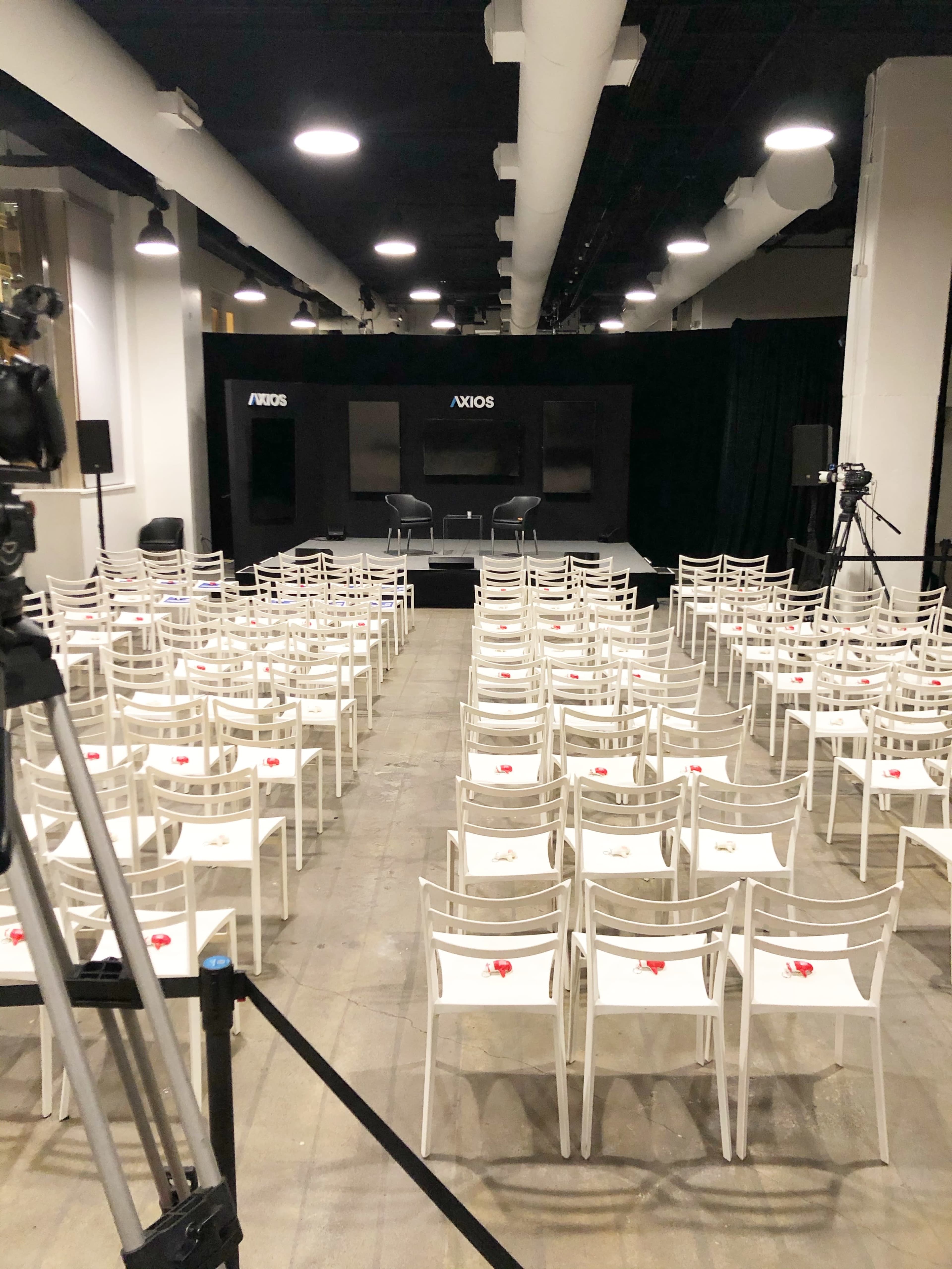 A spacious indoor venue is set up for an event, featuring rows of empty white chairs facing a stage with Axios branding and cameras positioned for recording.