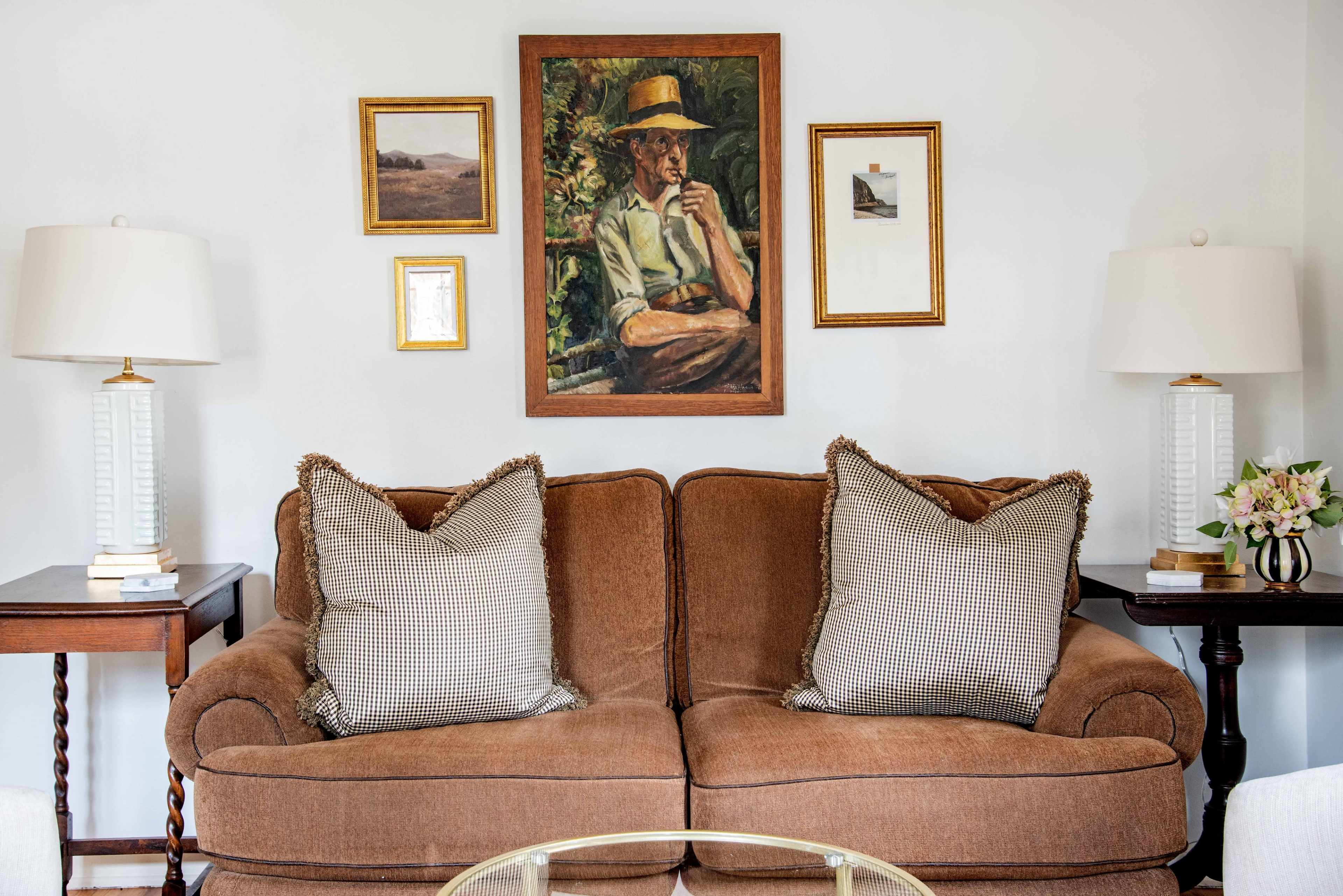 The image features a brown sofa with checkered pillows, flanked by two lamps, and a wall adorned with a portrait and framed pictures.