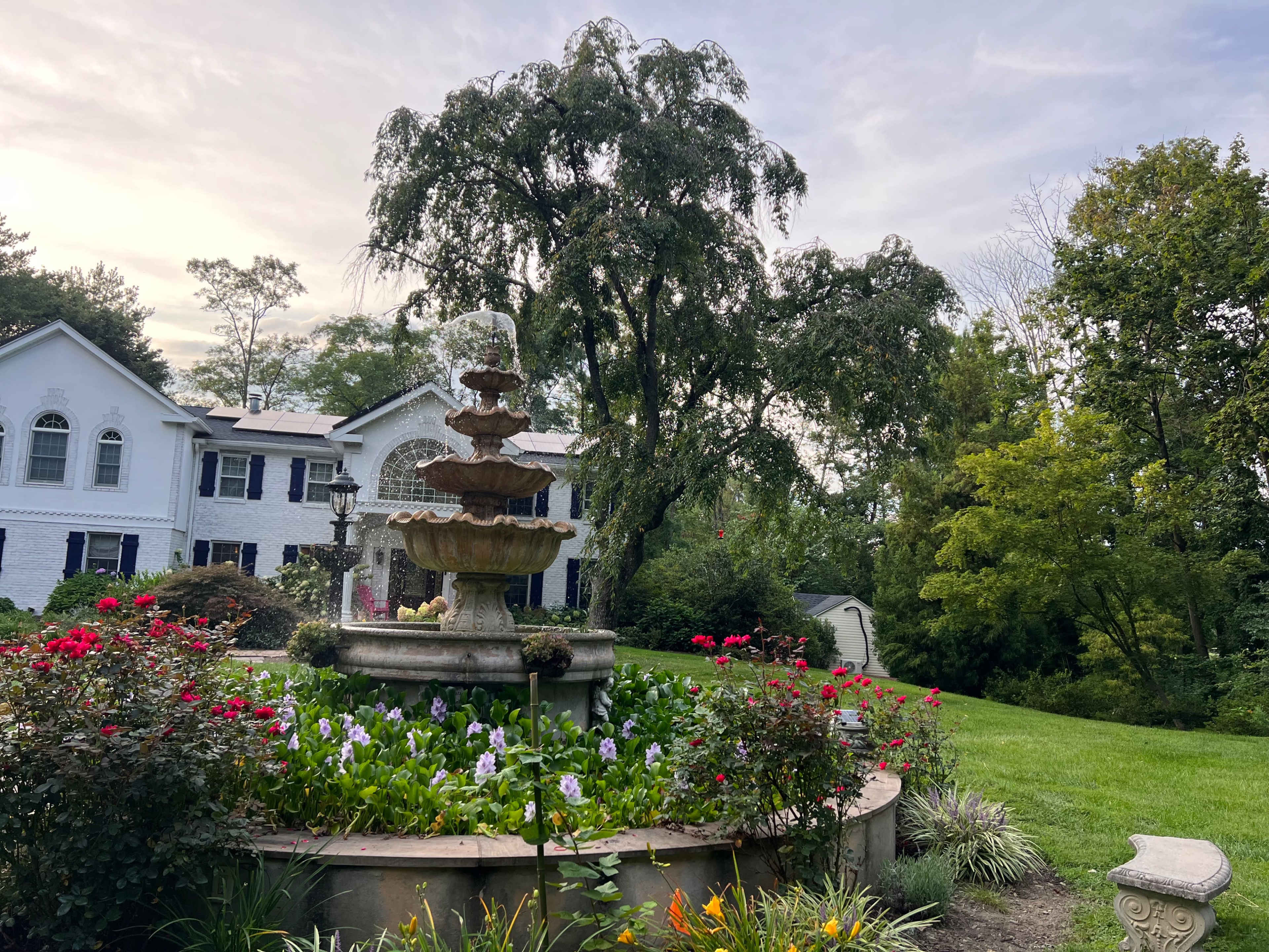 A landscaped garden with a fountain surrounded by colorful flowers, set in front of a large white house with blue shutters.