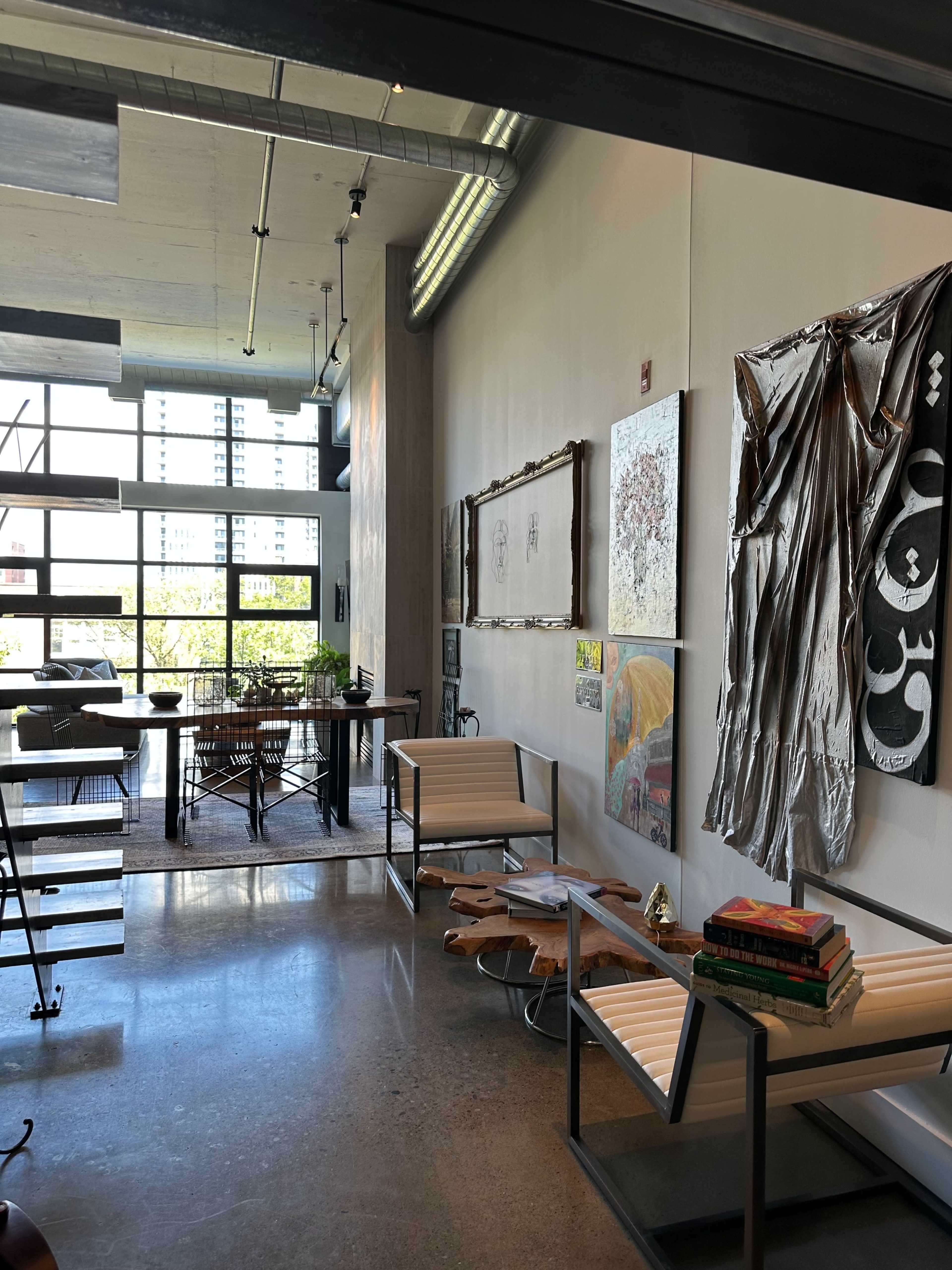 The image shows a modern office space featuring a mix of artwork, a dining area, and contemporary furniture against a backdrop of large windows.