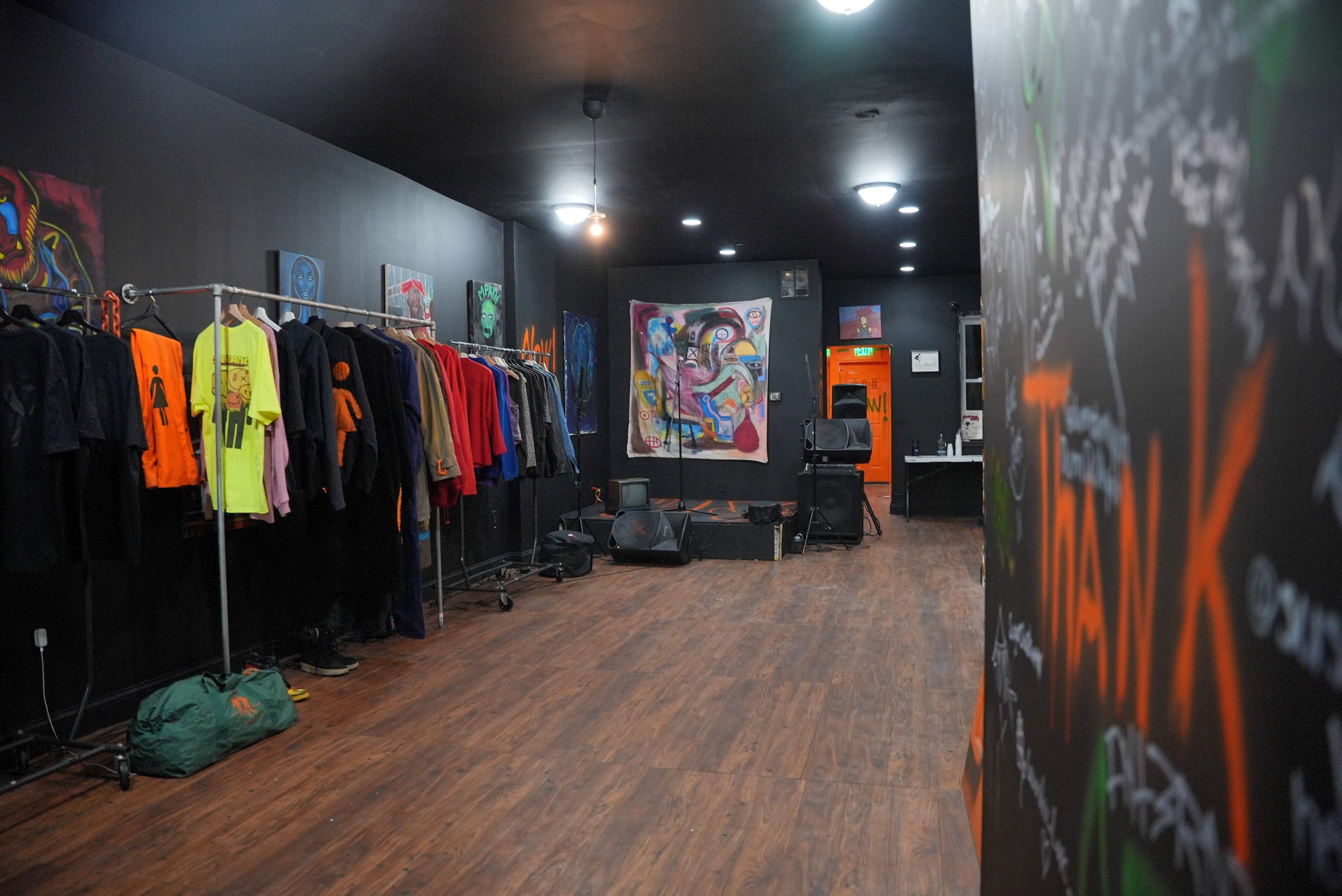 A clothing boutique with a rack of colorful apparel, an empty performance area, and walls adorned with colorful artwork and writings.