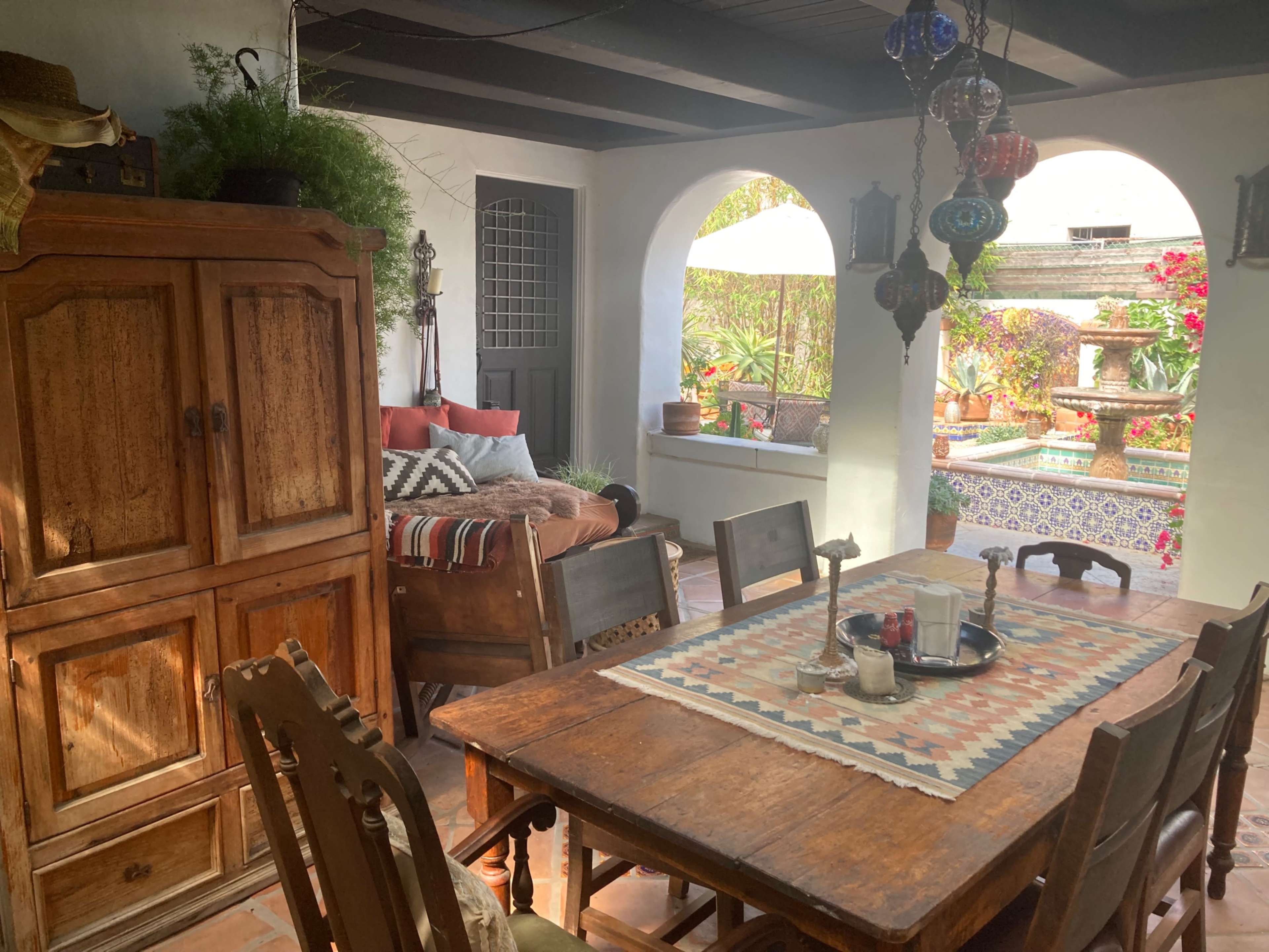 A dining area with a wooden table, surrounded by chairs, a carved wooden cabinet, and arches leading to a patio filled with plants and decorative tiles.
