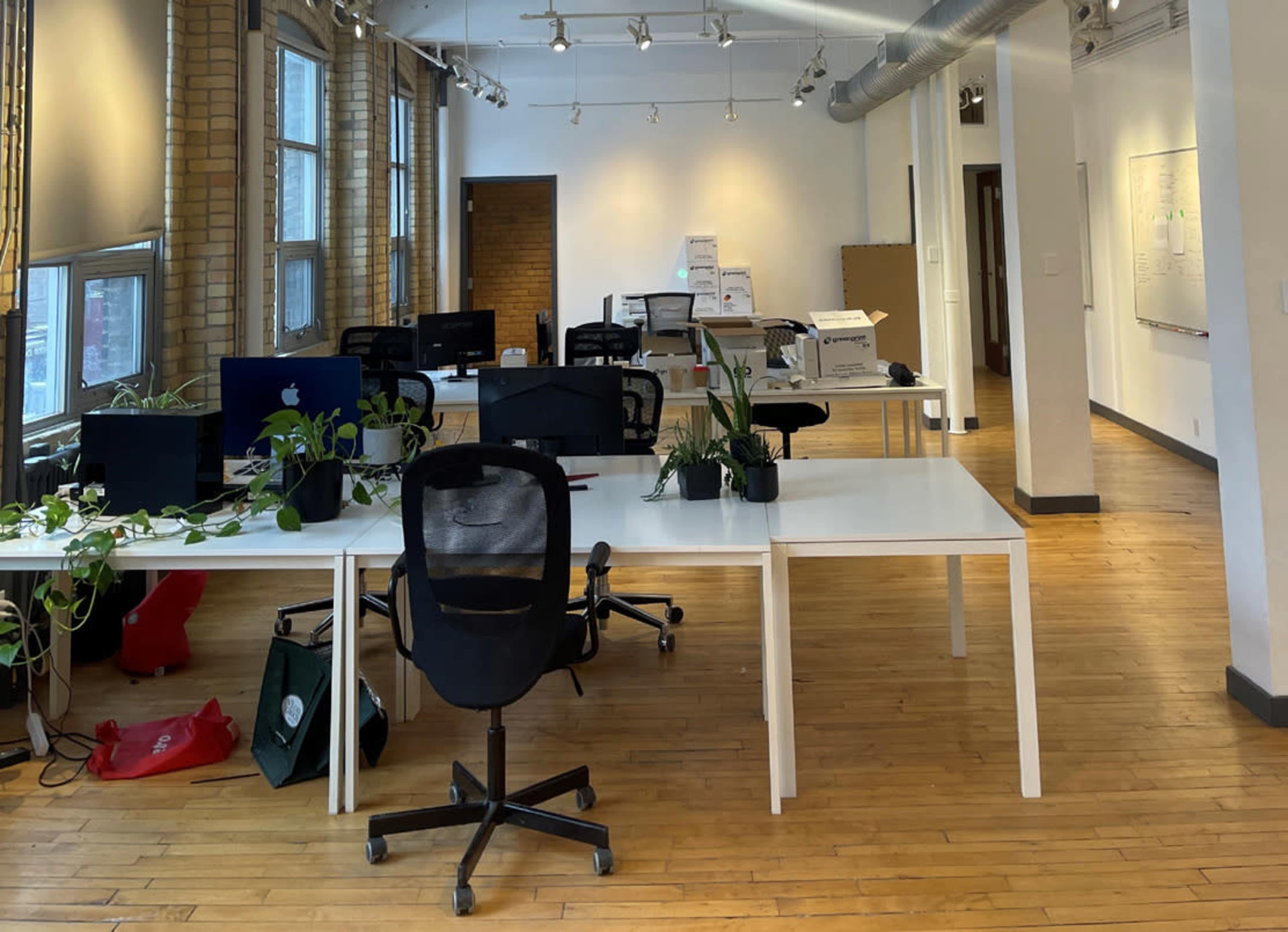 An office space with several white desks, rolling chairs, and minimal decorations, illuminated by overhead lighting.