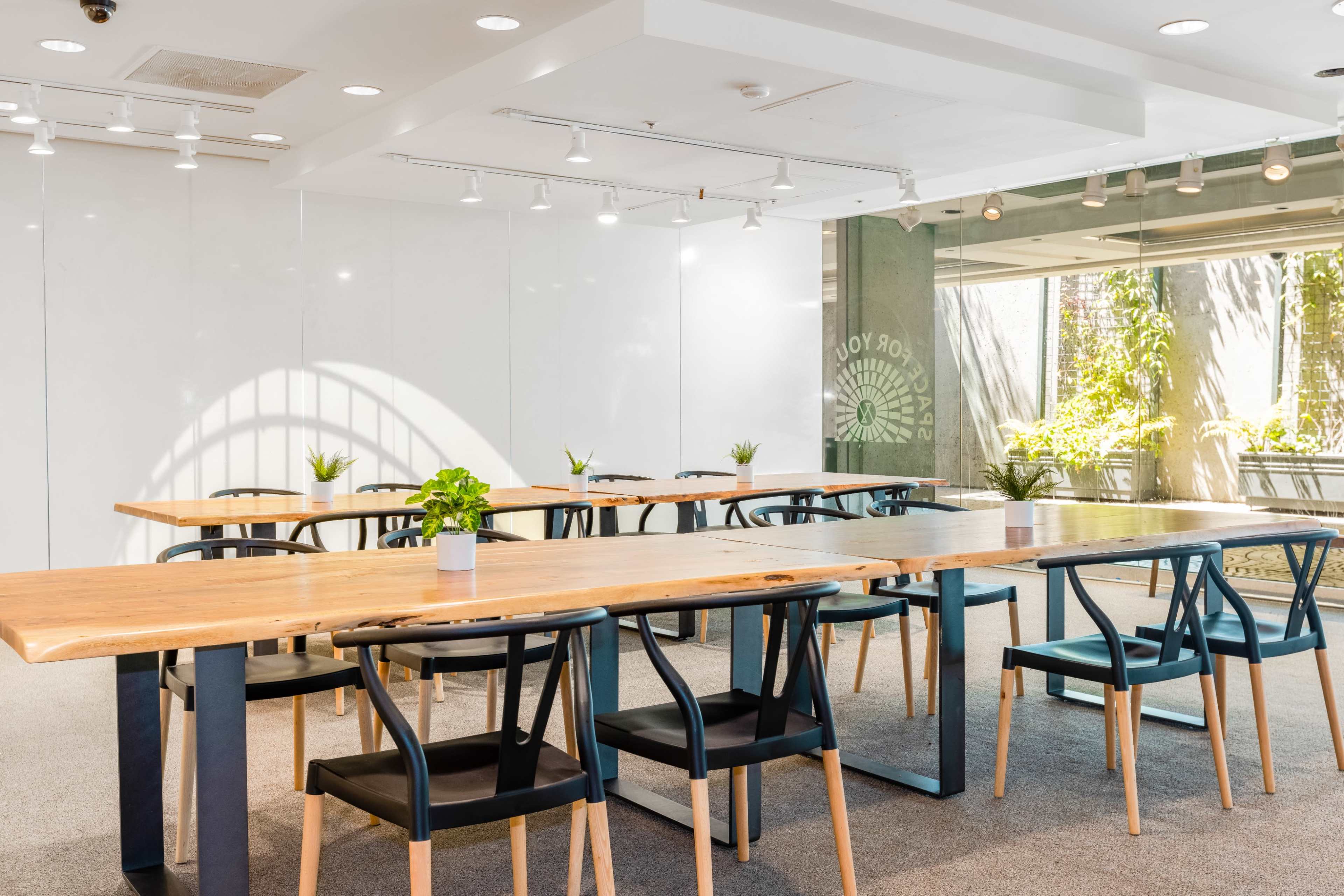 A large, well-lit meeting room features several long wooden tables with black chairs arranged neatly, surrounded by windows showcasing greenery outside.