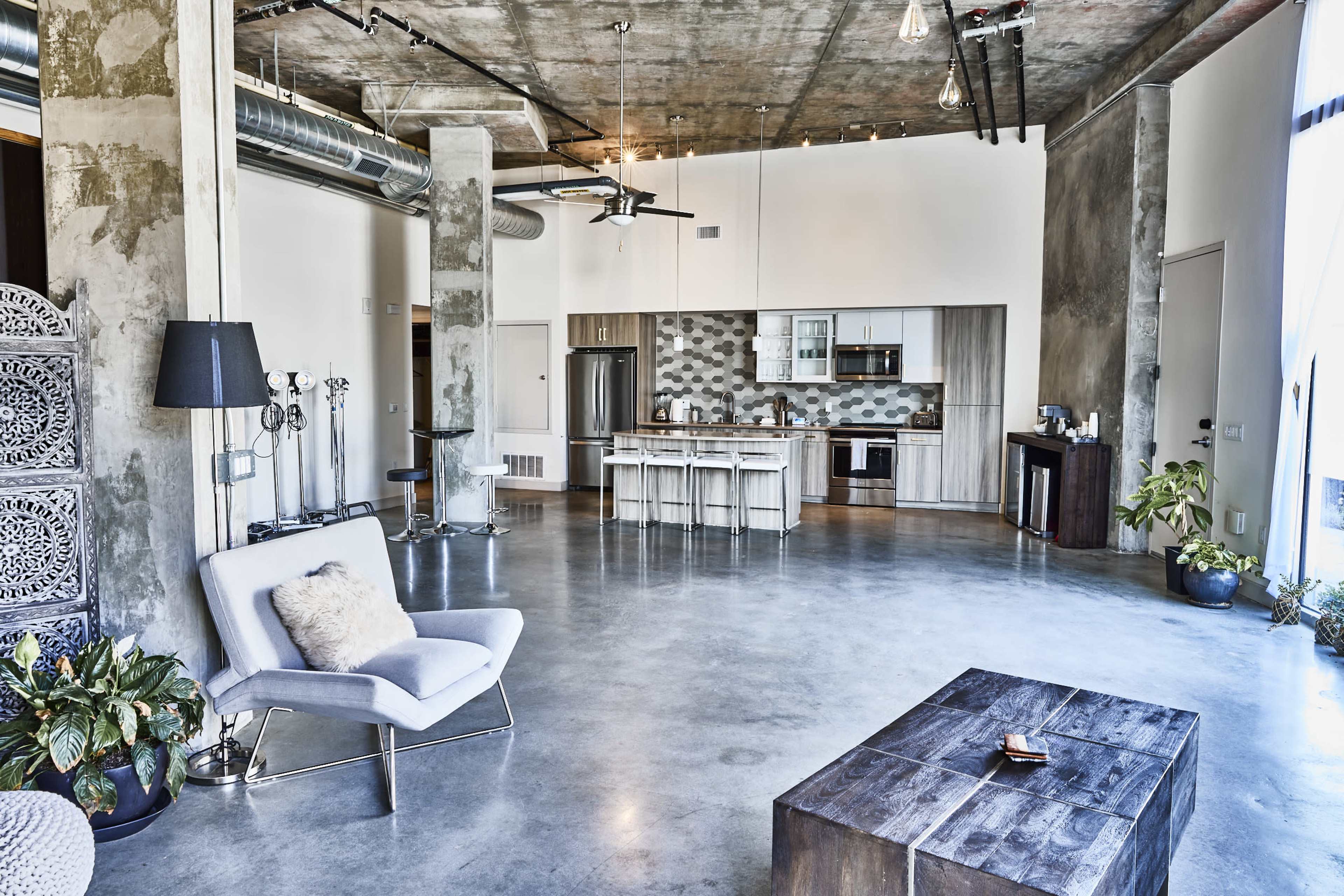 A spacious modern loft features a concrete floor, an open kitchen with wood cabinetry, and a seating area with minimalist furniture.