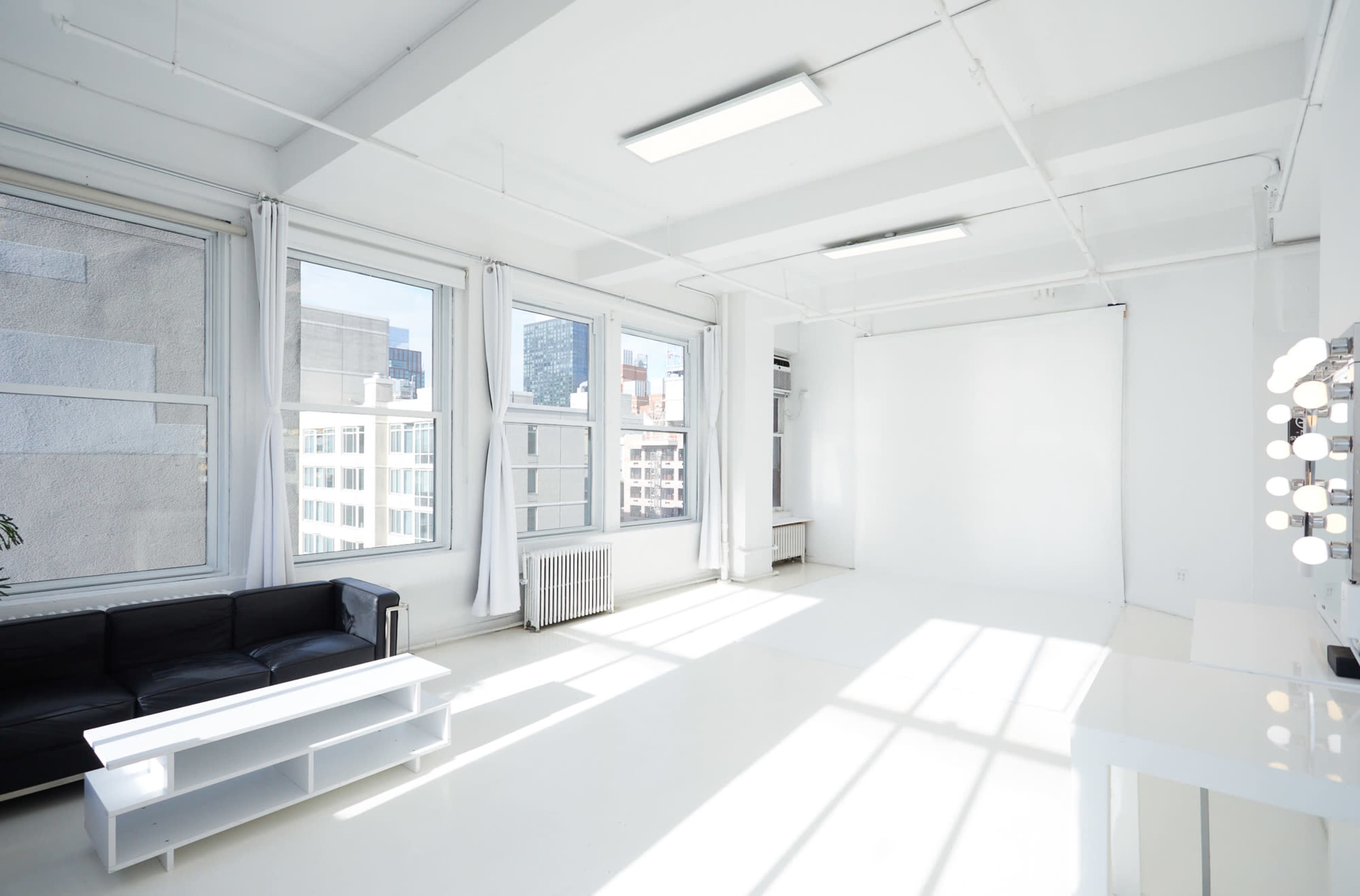 A bright, minimalist studio space features large windows, white walls, and a black sofa with a small coffee table.