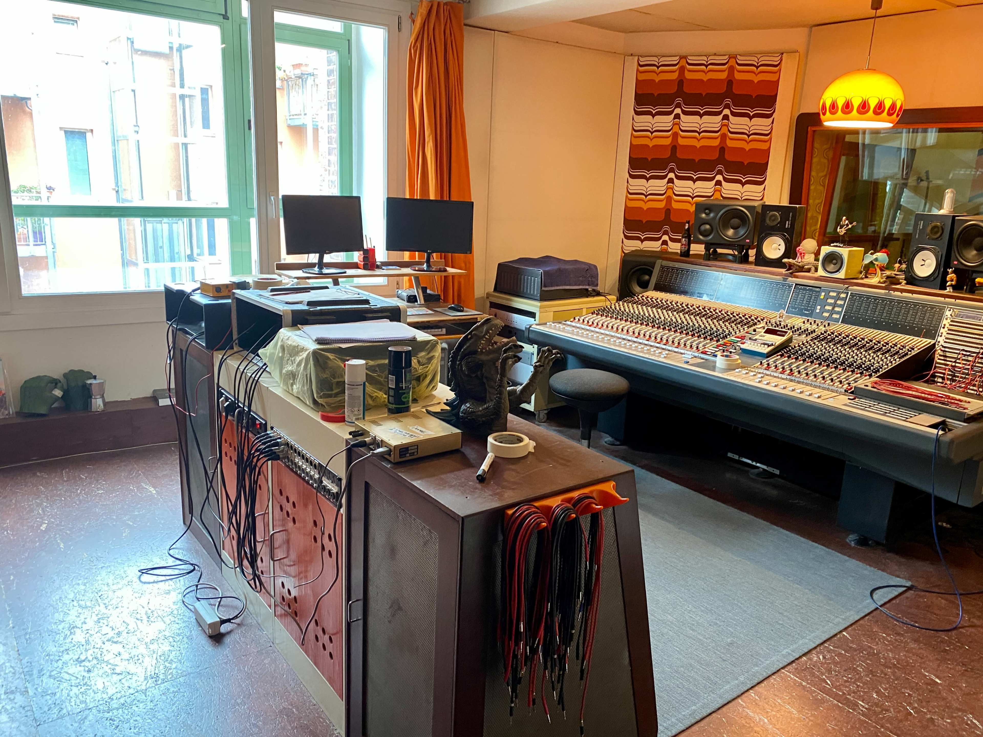A music studio with a large mixing console, computer monitors, and various equipment arranged on a desk, surrounded by a colorful patterned wall.