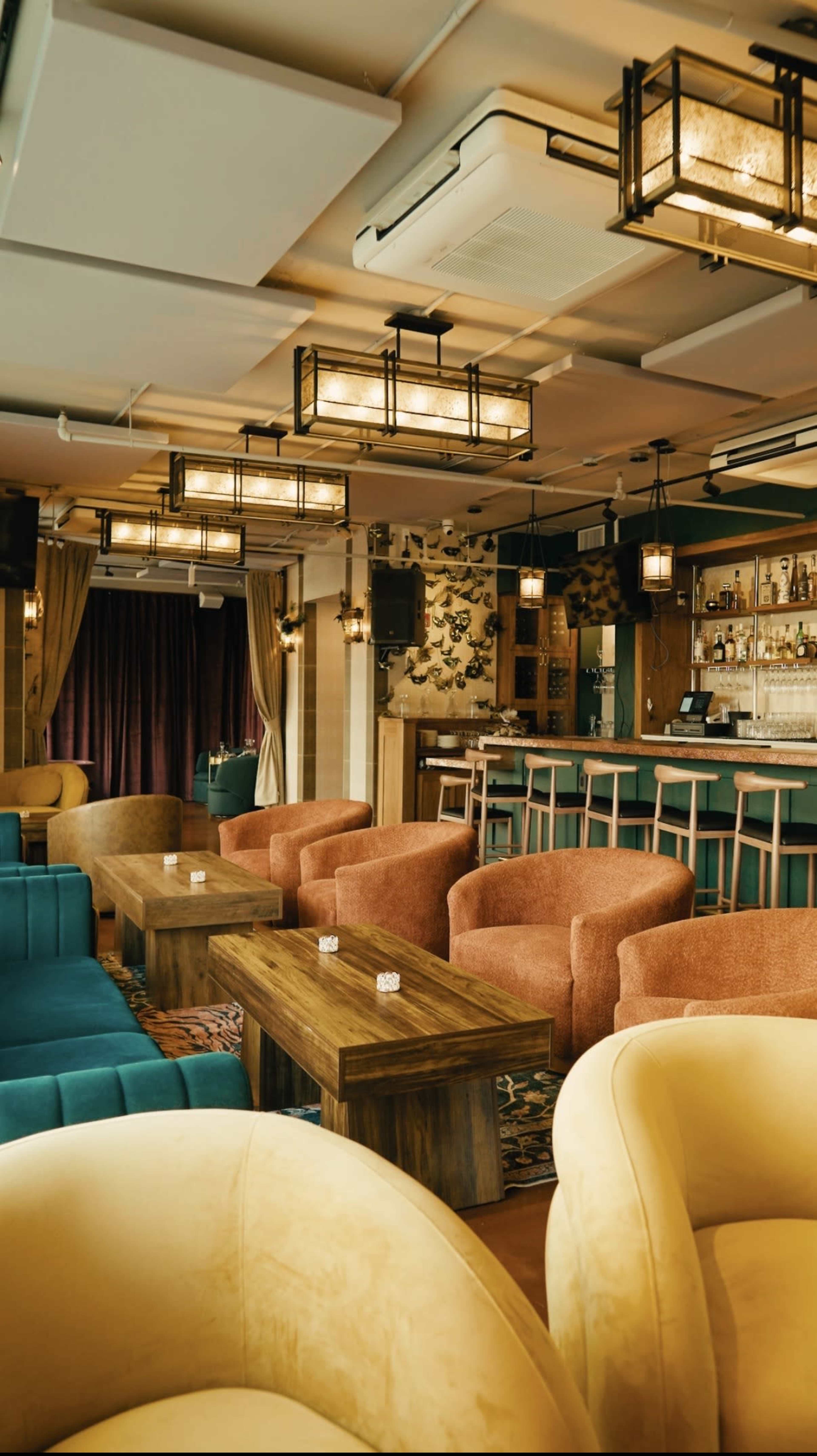 A lounge area features colorful upholstered chairs and wooden tables arranged in front of a bar with decorative lighting and a backdrop of shelves.