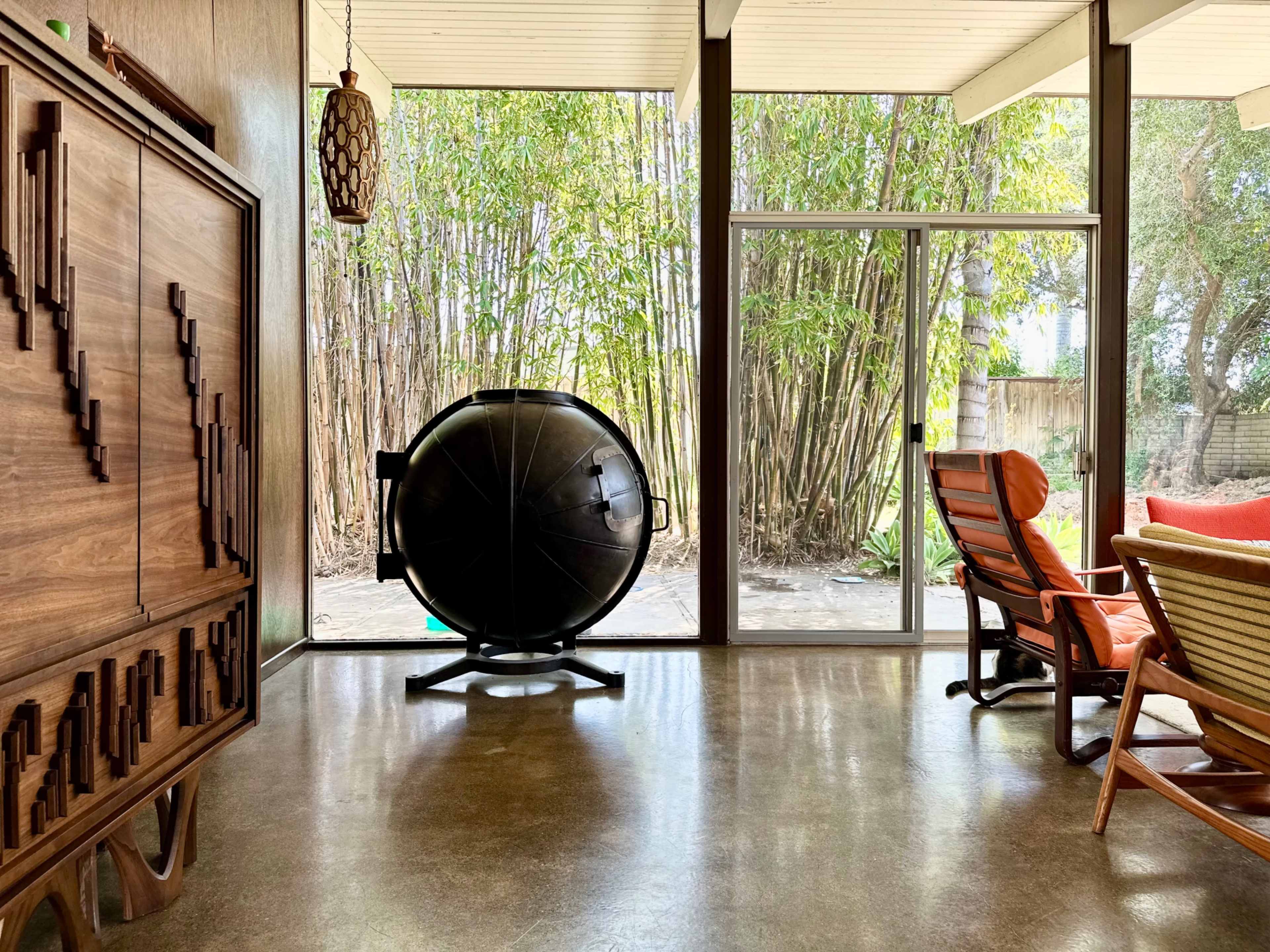 A modern living space features a round black object on a stand, with large glass windows revealing a view of bamboo plants outside.