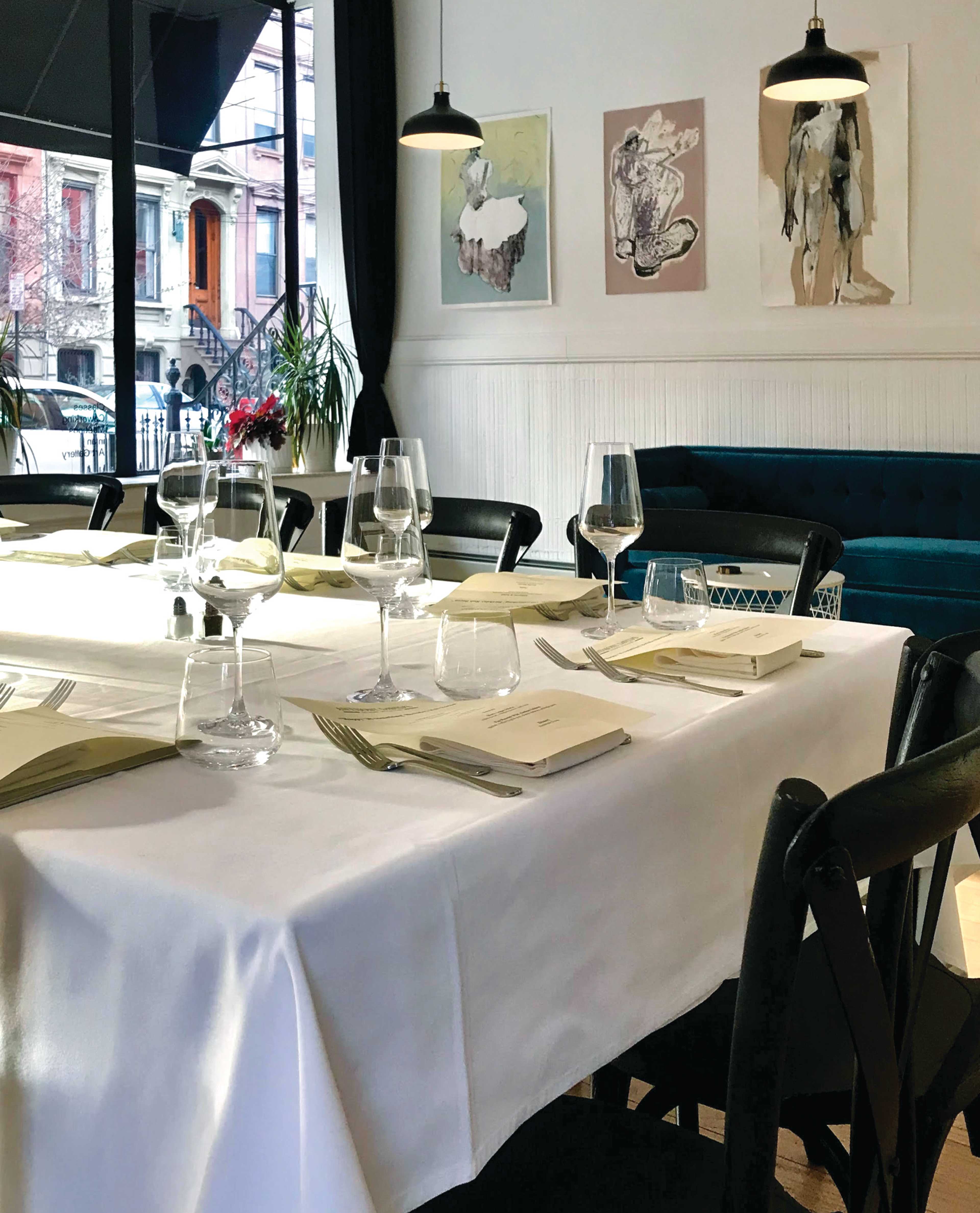 A dining table is set with glasses and utensils, and the room features artwork on the walls and a view of the street through large windows.