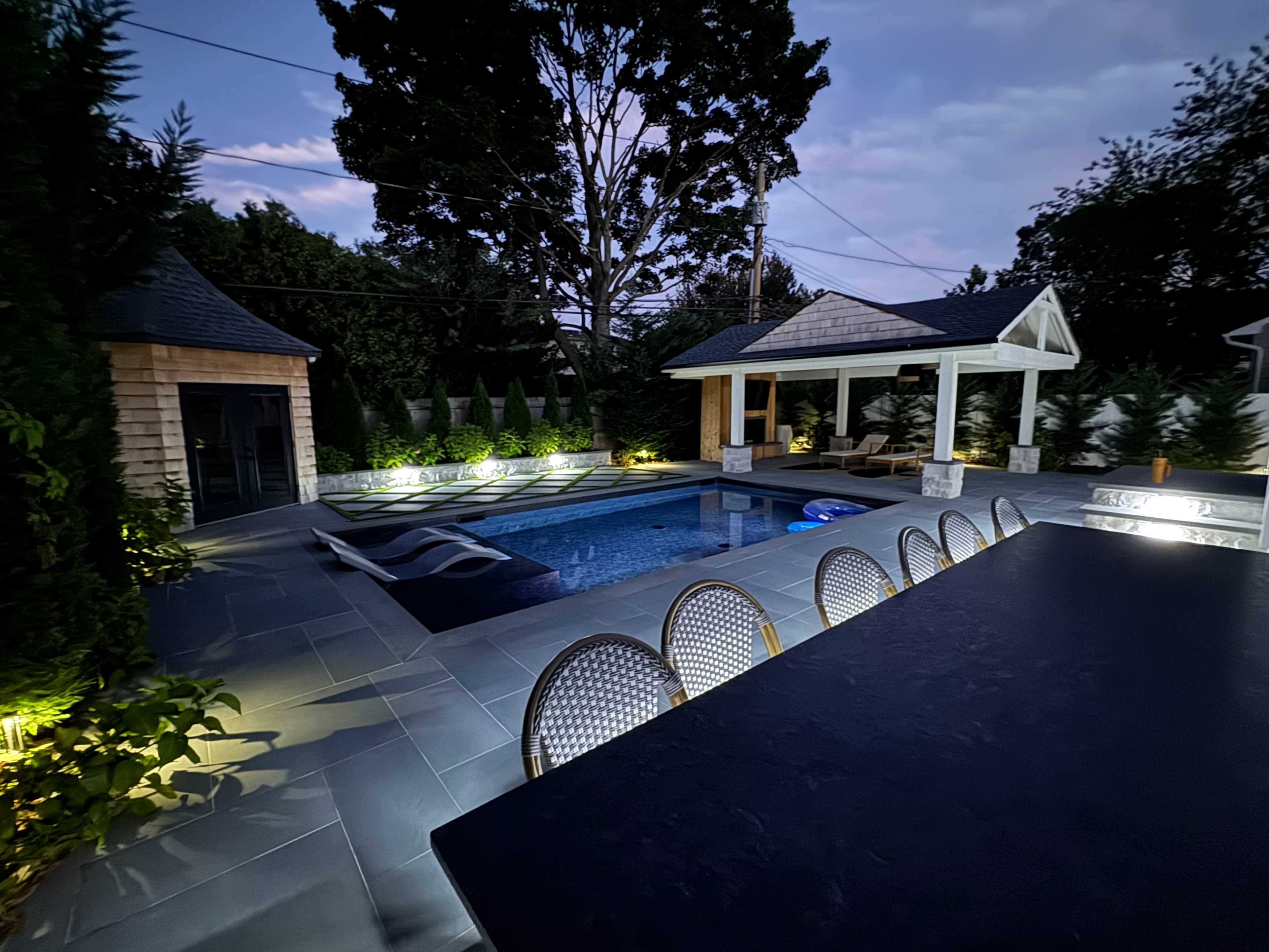 The image shows a modern outdoor space featuring a swimming pool, lounge chairs, and a covered seating area, all illuminated by soft lighting in the evening.