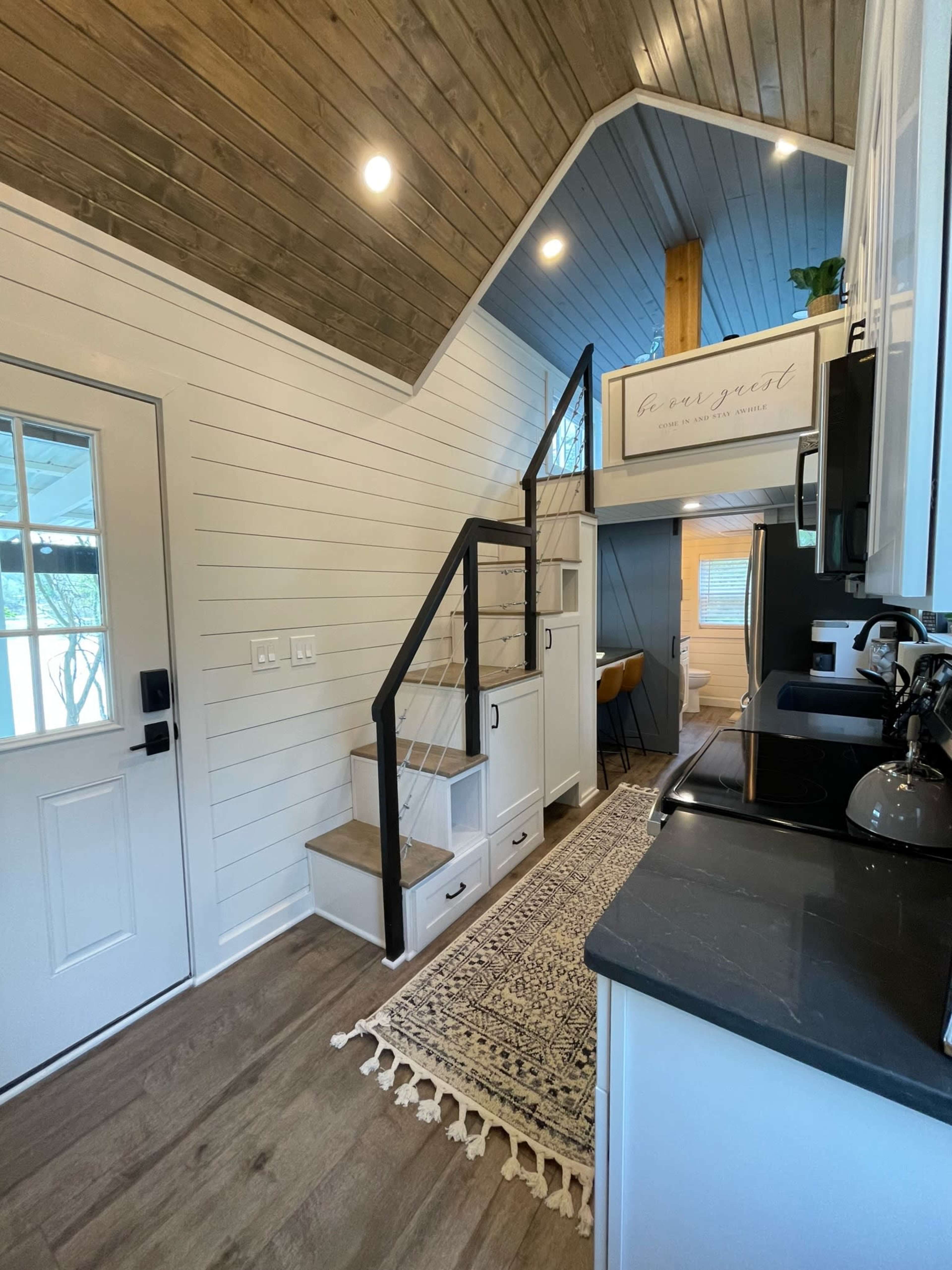 The interior of a small house featuring a stairway, modern kitchen elements, and a welcoming entrance with a decorative rug.