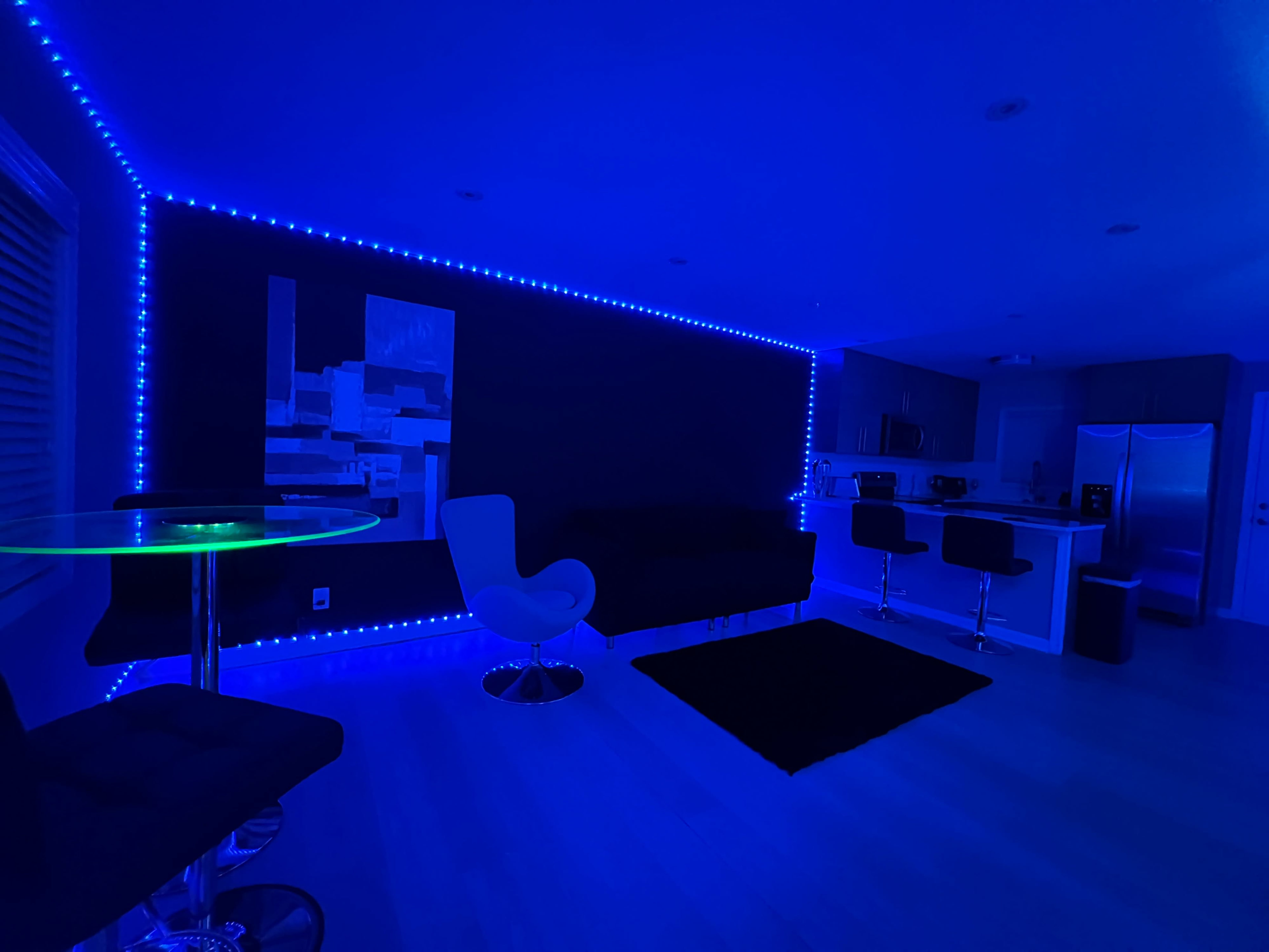 The scene features a dimly lit modern living space with blue LED lights outlining the walls and a sleek furniture setup including a dining table and bar stools.