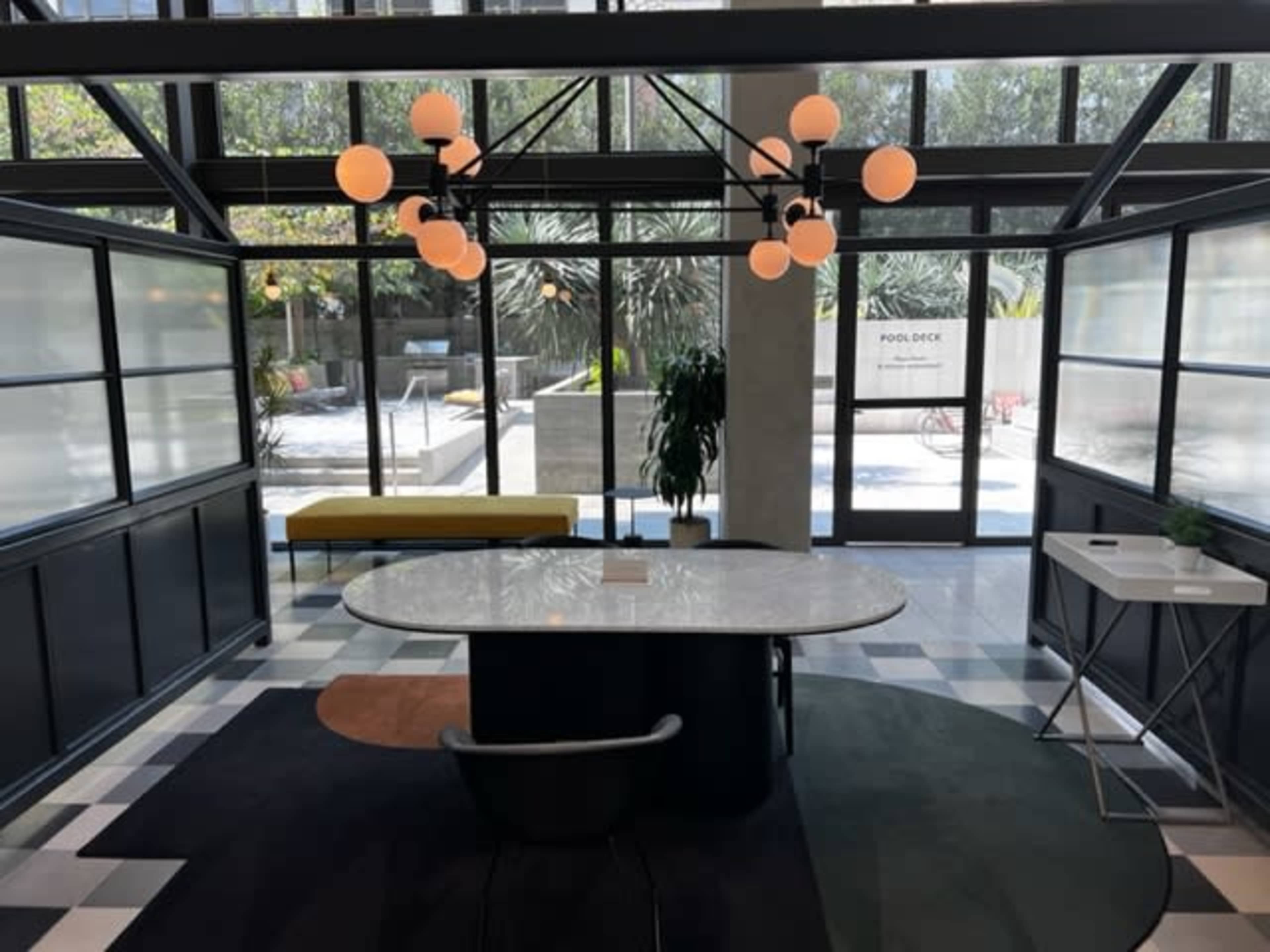 The image shows a modern interior space with a circular marble table, a black chair, a green bench, and large windows overlooking a landscaped outdoor area.
