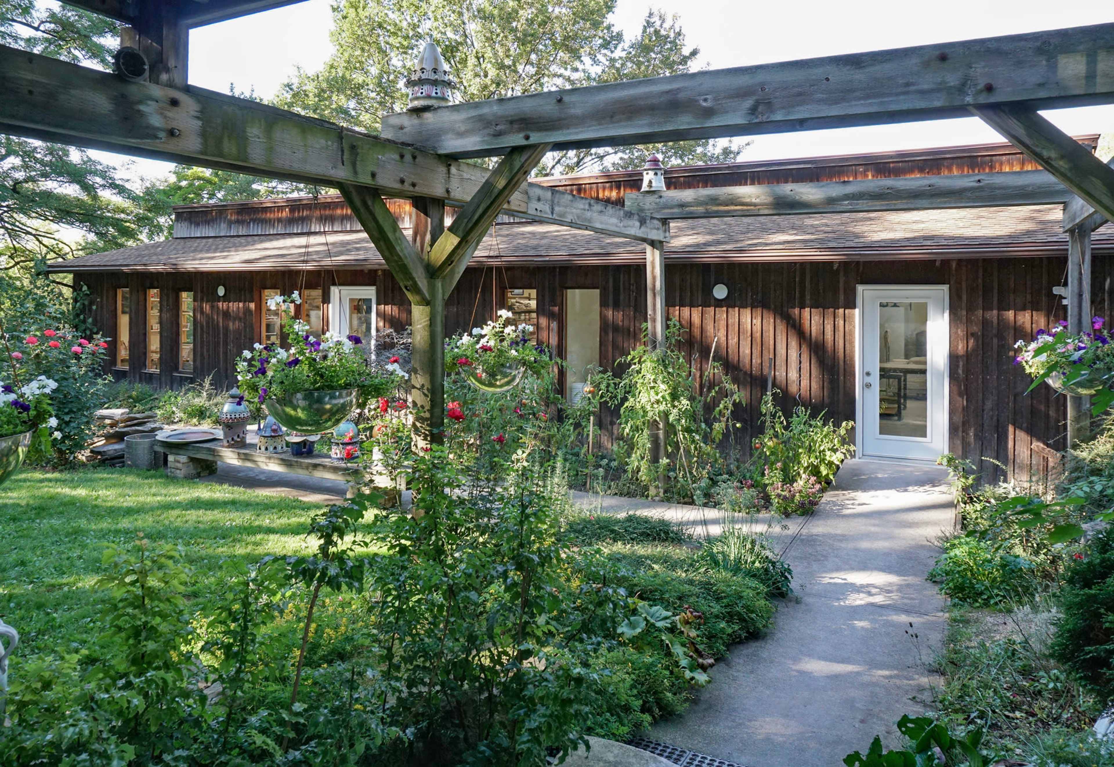 The image shows a wooden single-story house surrounded by a garden with blooming flowers and a stone pathway leading to the entrance.
