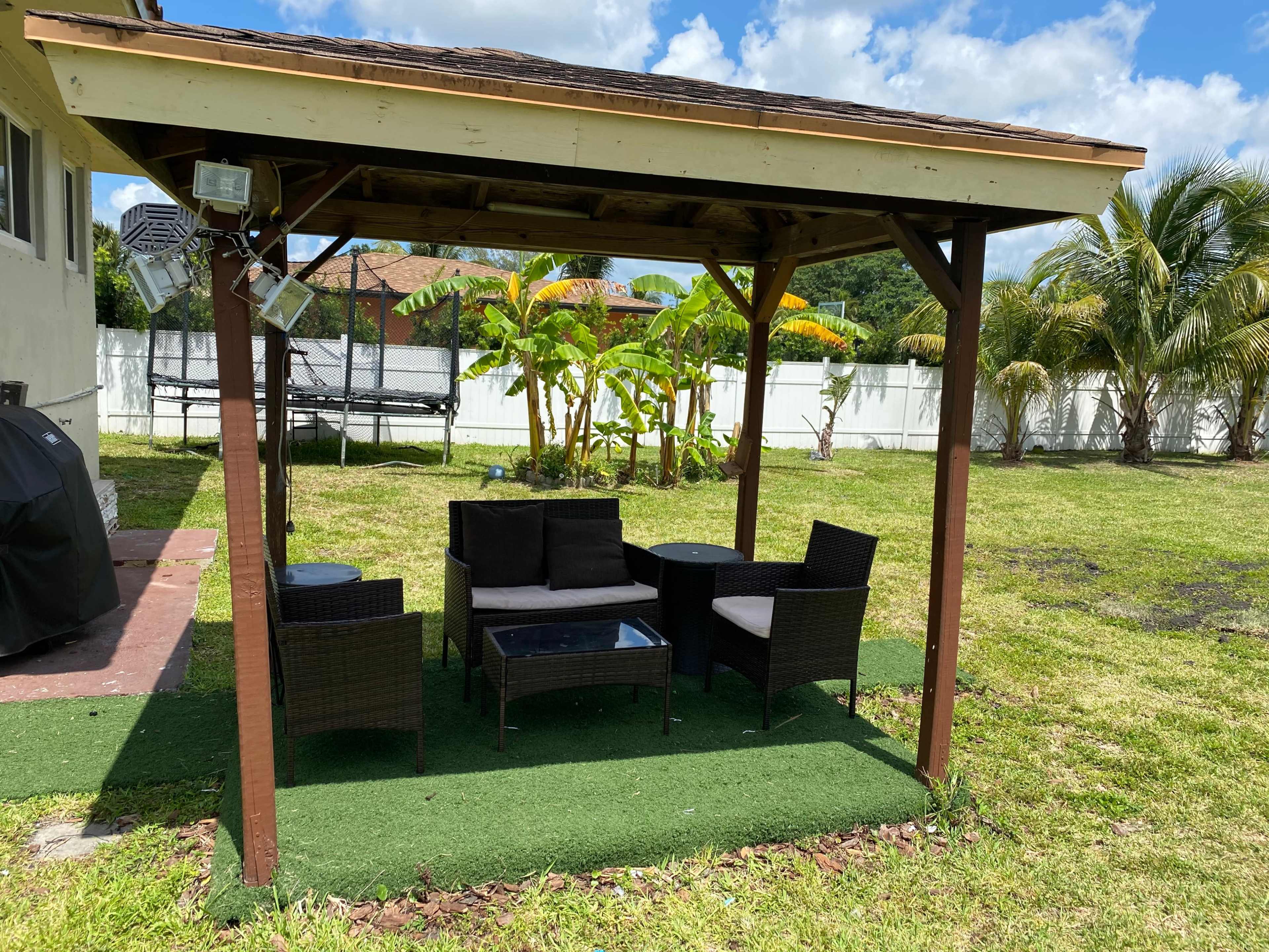 A shaded patio with a seating area made of wicker furniture and a glass table is set on green grass in a backyard surrounded by palm trees and a white fence.