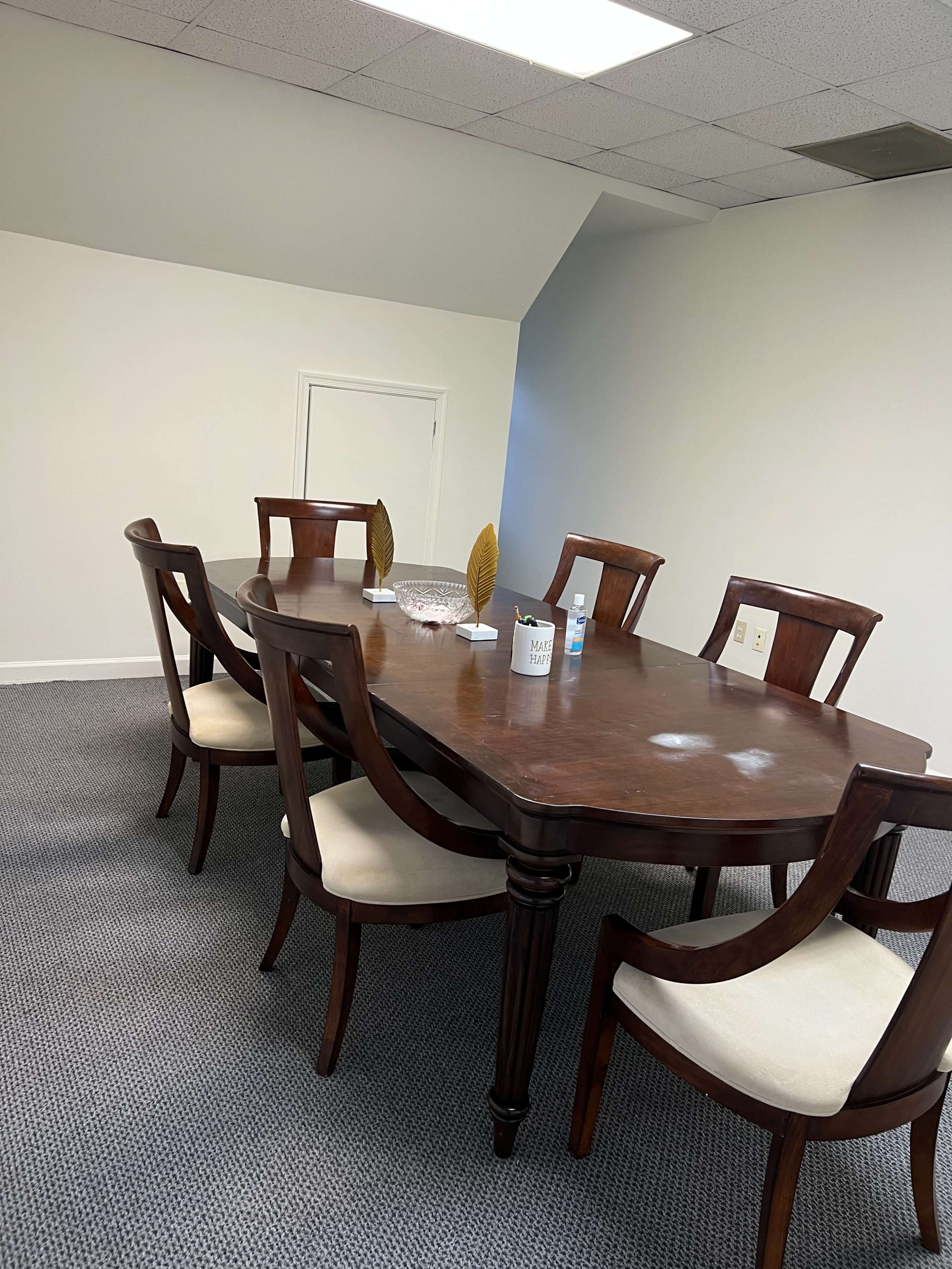 A wooden conference table is surrounded by eight upholstered chairs in a sparsely furnished room.