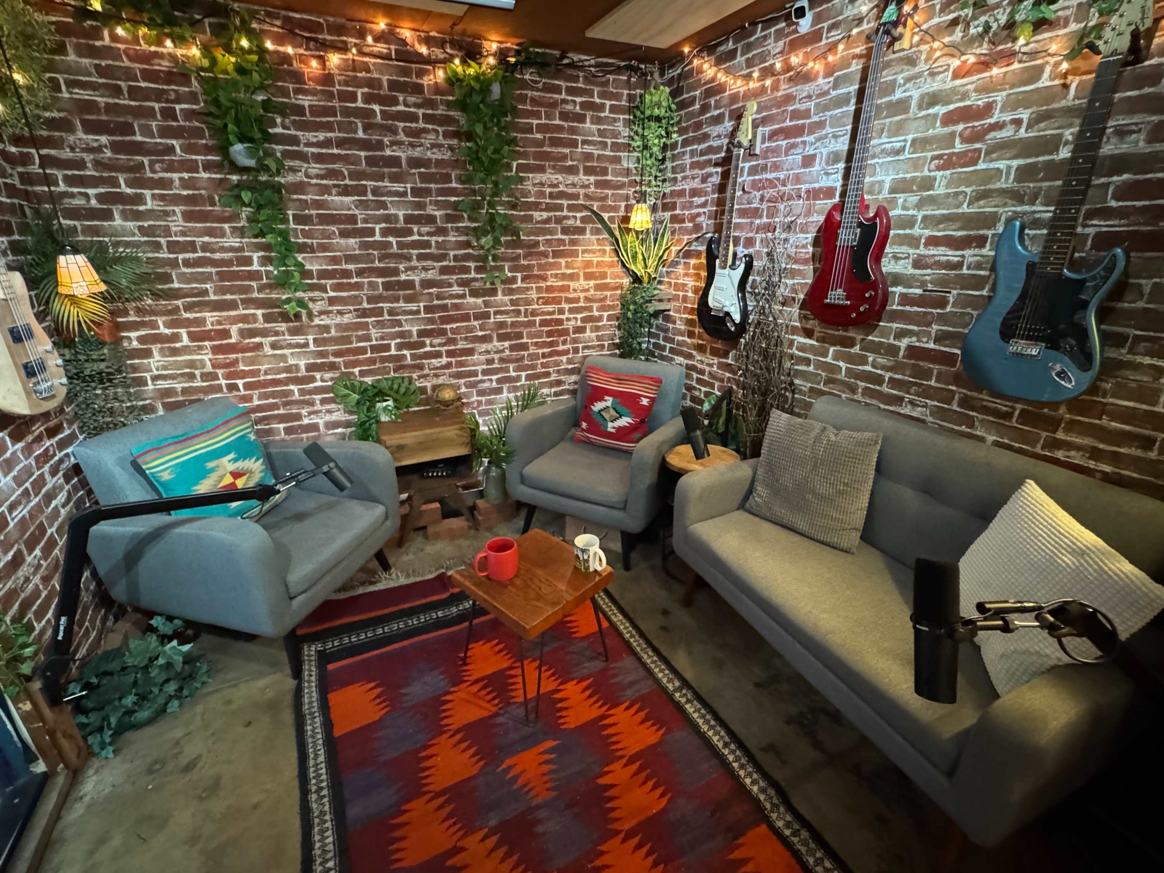 The image shows a cozy lounge with brick walls, two gray sofas, a table with mugs, and guitars hanging on the wall amidst decorative plants.