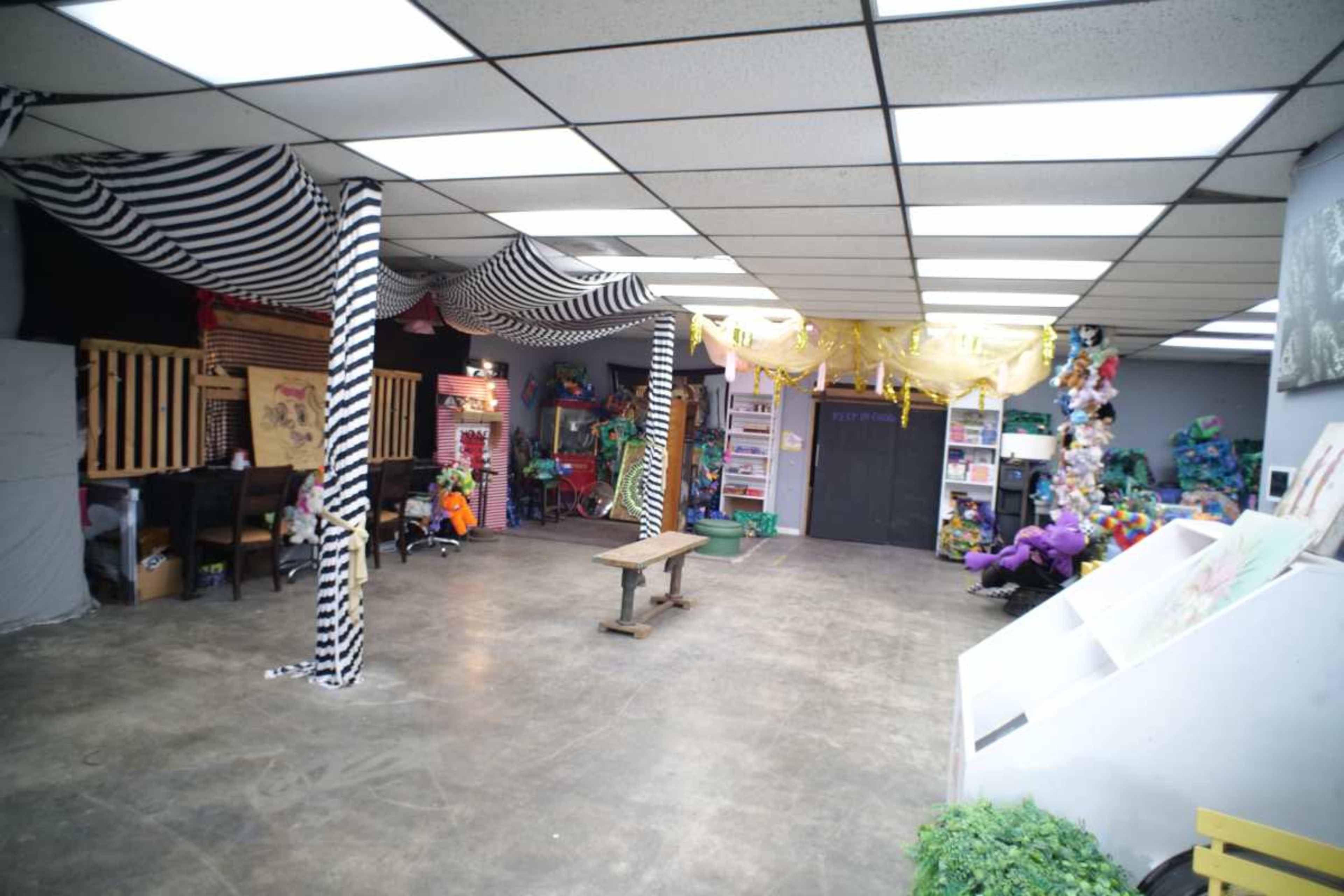The image shows a large, open room with a concrete floor, decorated with various colorful items and draped fabrics, and featuring storage shelves against the walls.