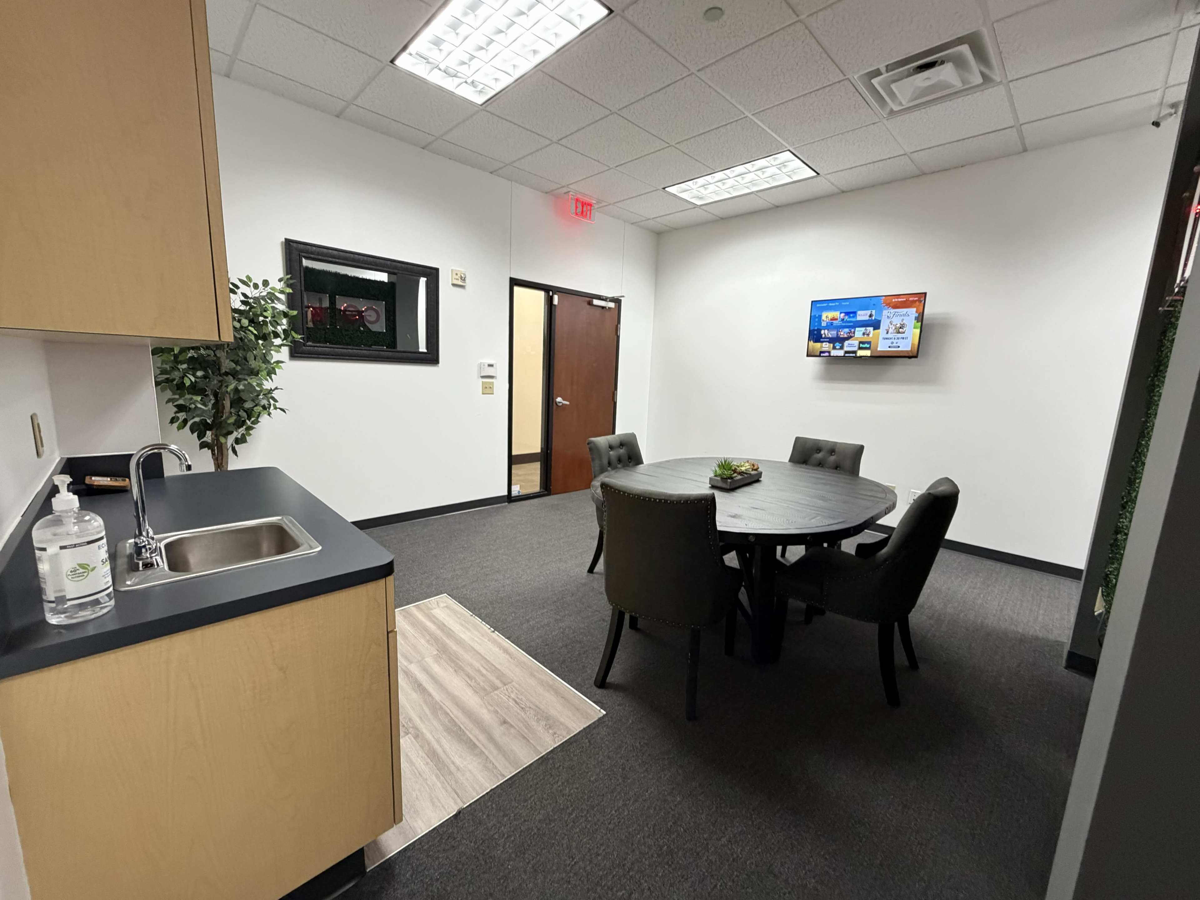The image shows a small meeting room with a round table and chairs, a kitchenette area, and a wall-mounted screen.