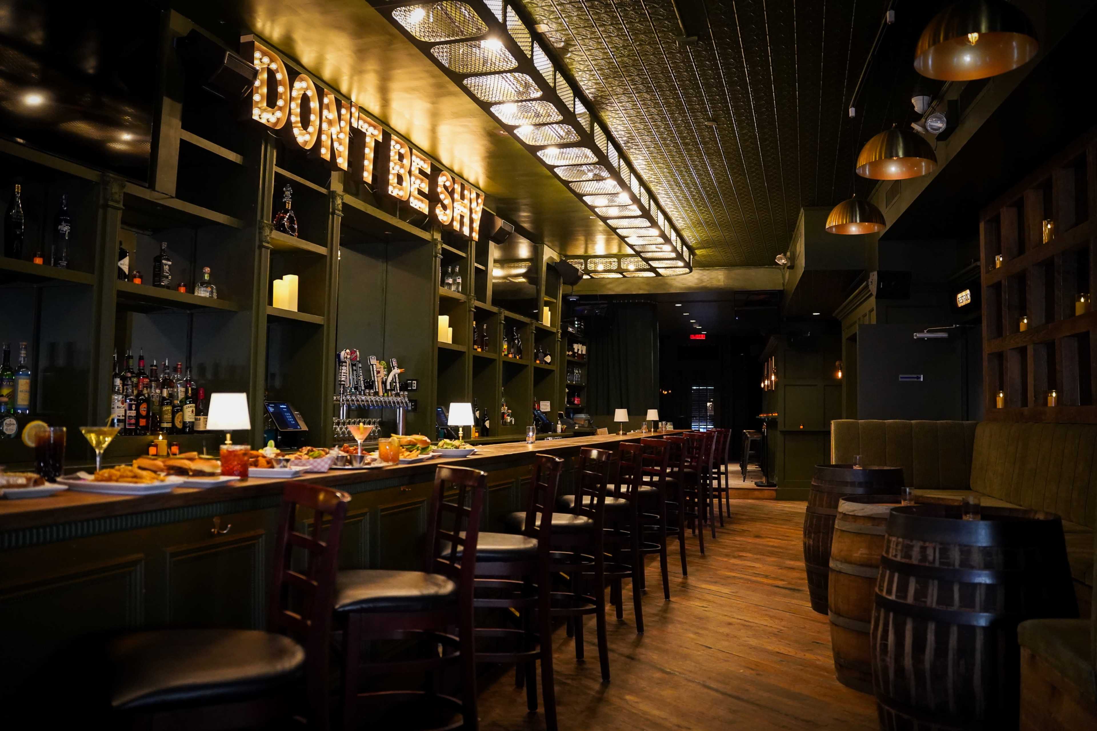 The image shows a bar with wooden stools, a long countertop lined with food and drinks, and a sign above the bar that reads "DON'T BE SHY."
