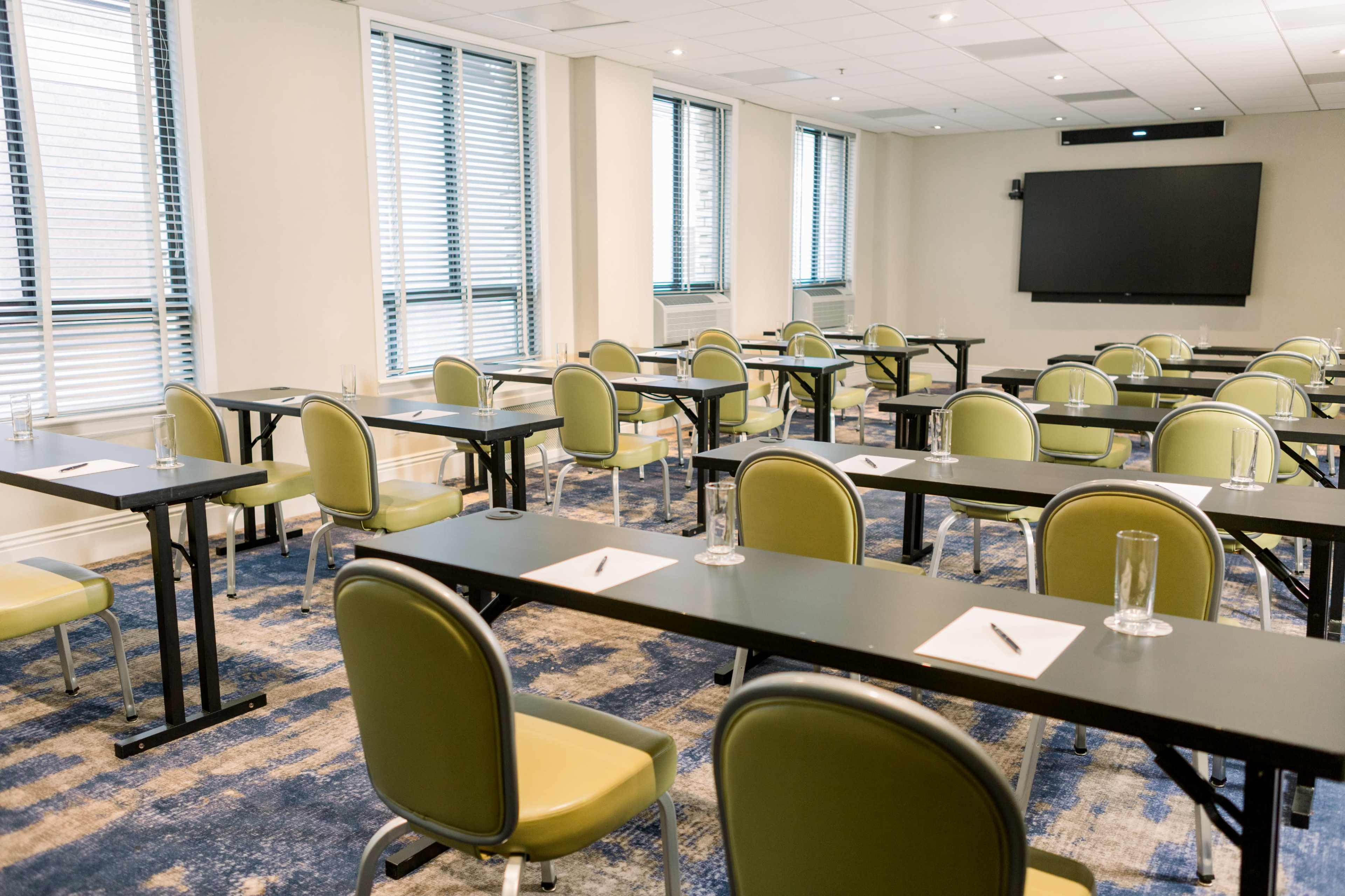 A classroom is arranged with black tables and green chairs, featuring notepads and pens at each seating station, and large windows along one wall.