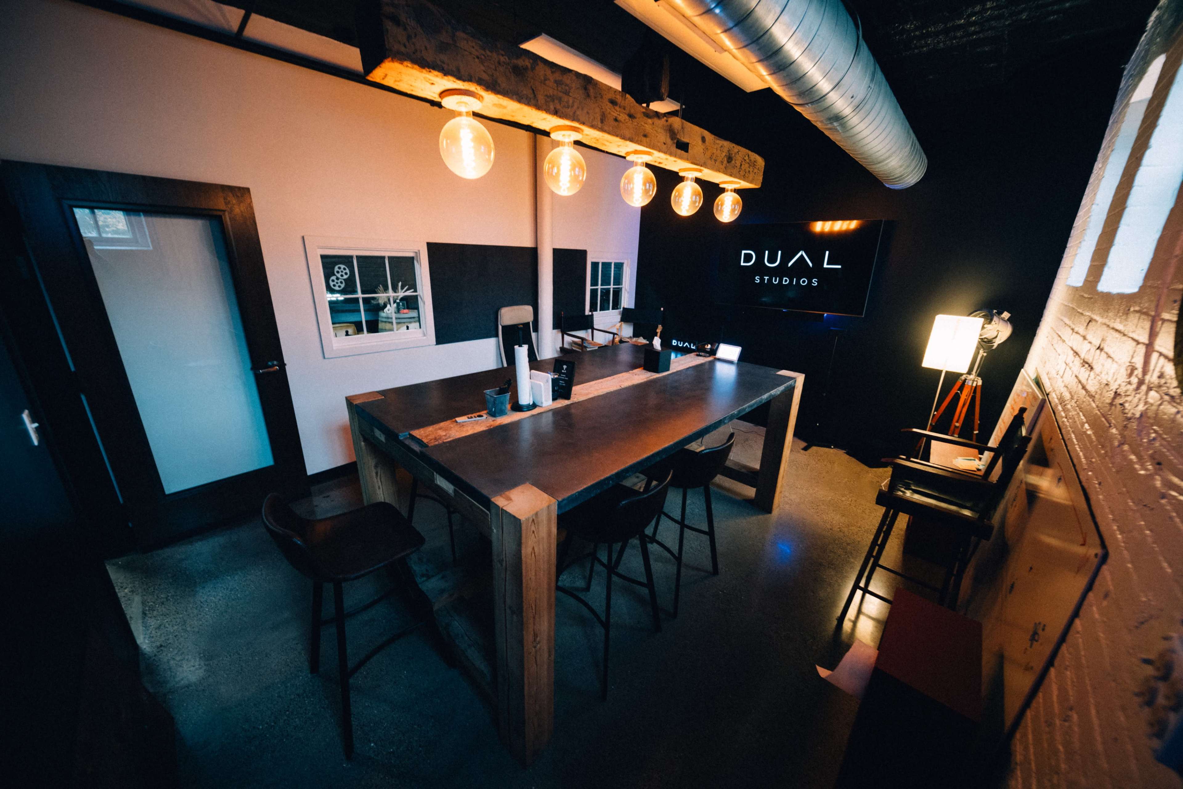 A modern office space features a large wooden table surrounded by chairs, with a television displaying the "DUAL Studios" logo and industrial lighting overhead.