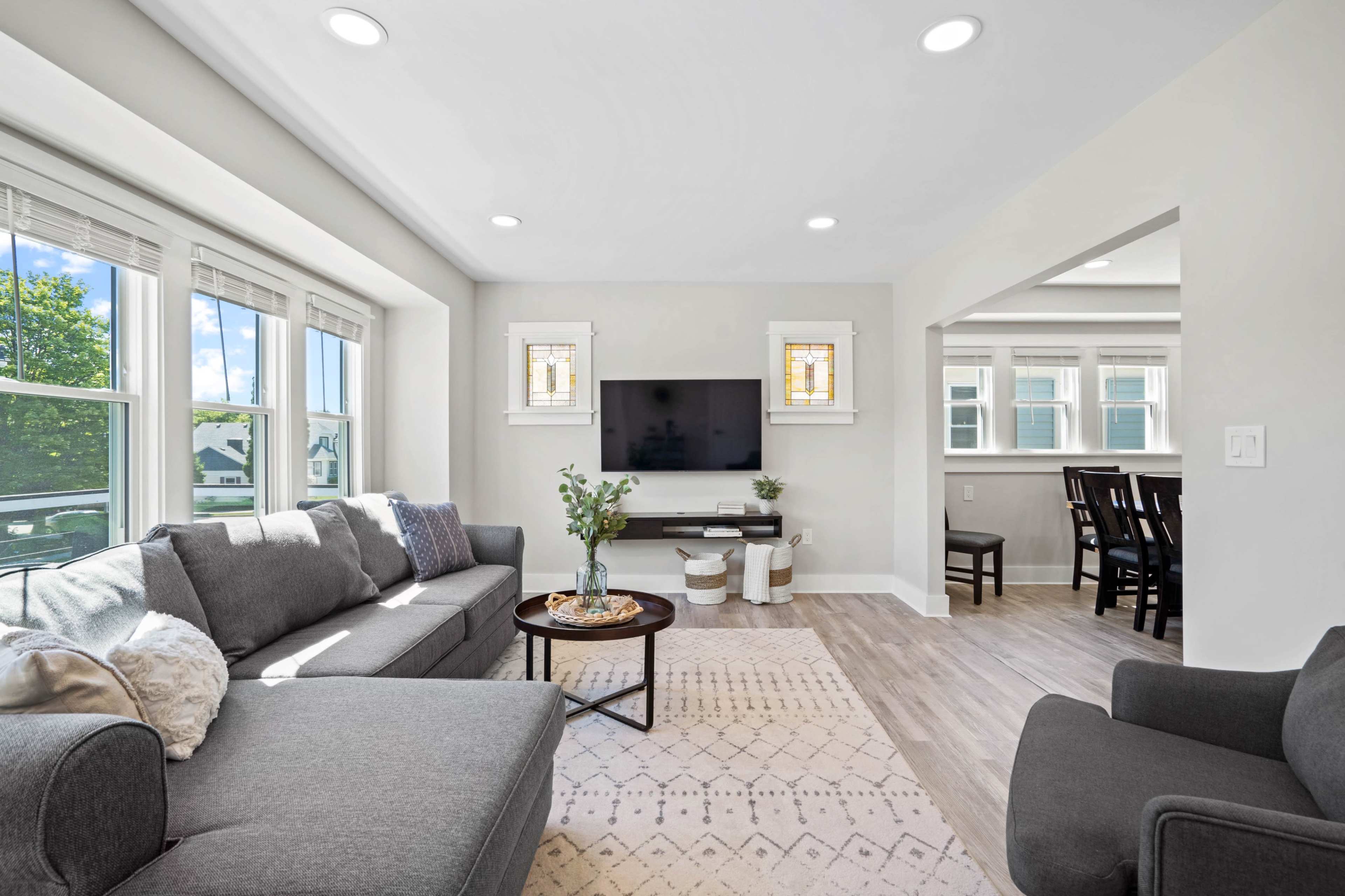A bright living room with a gray sectional couch, a coffee table, a mounted TV, and a dining area visible in the adjoining space.