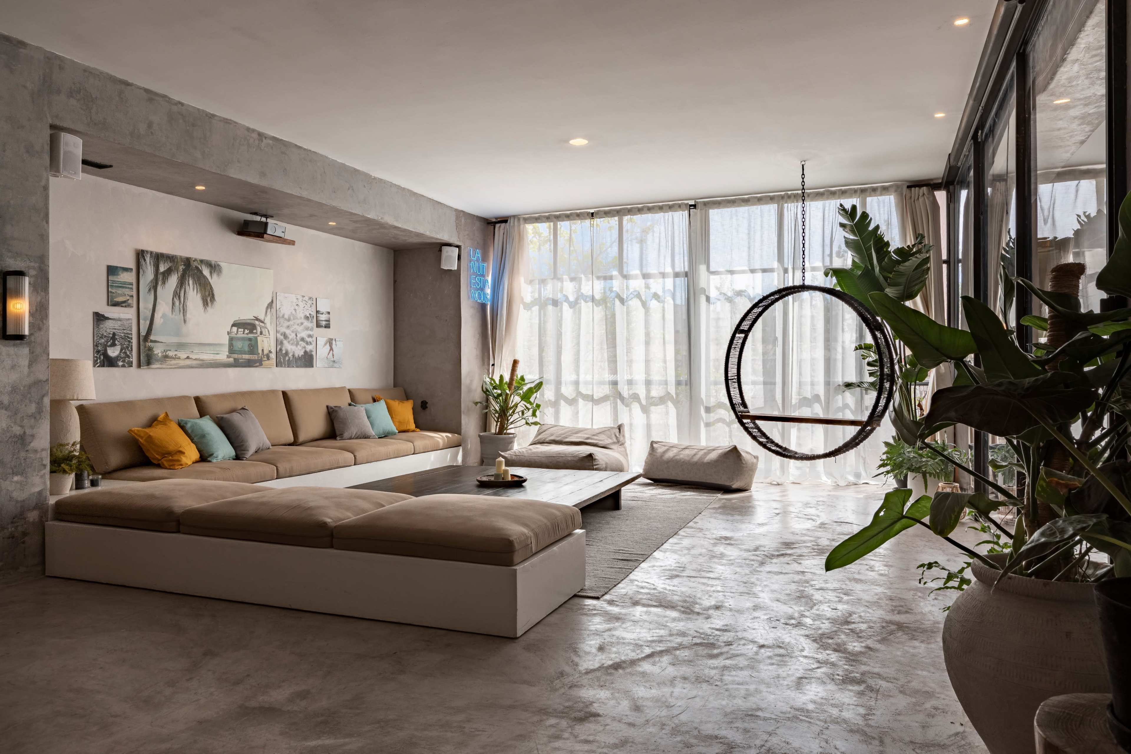A spacious living room features a large beige sectional sofa with colorful throw pillows, large windows with sheer curtains, and a circular hanging chair.