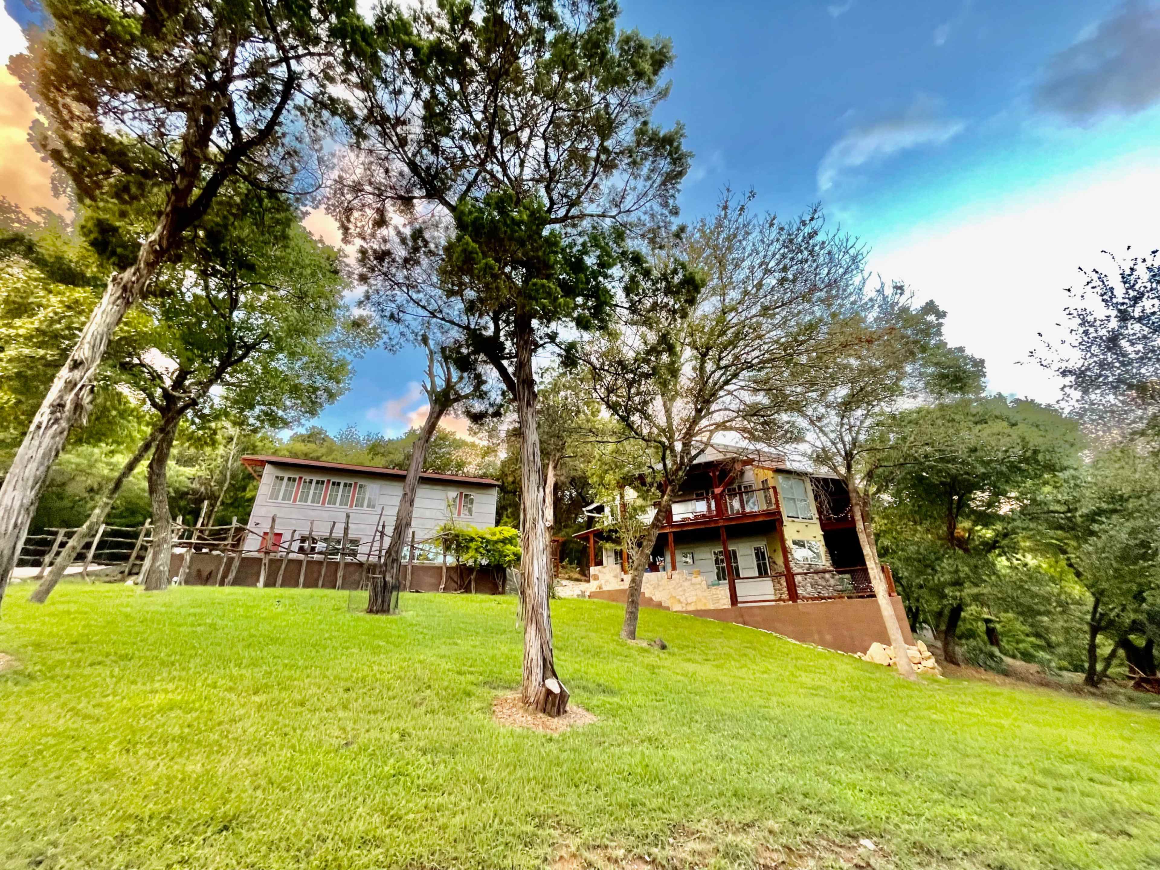 A two-story house with a deck and a nearby single-story structure is situated on a grassy slope surrounded by trees.