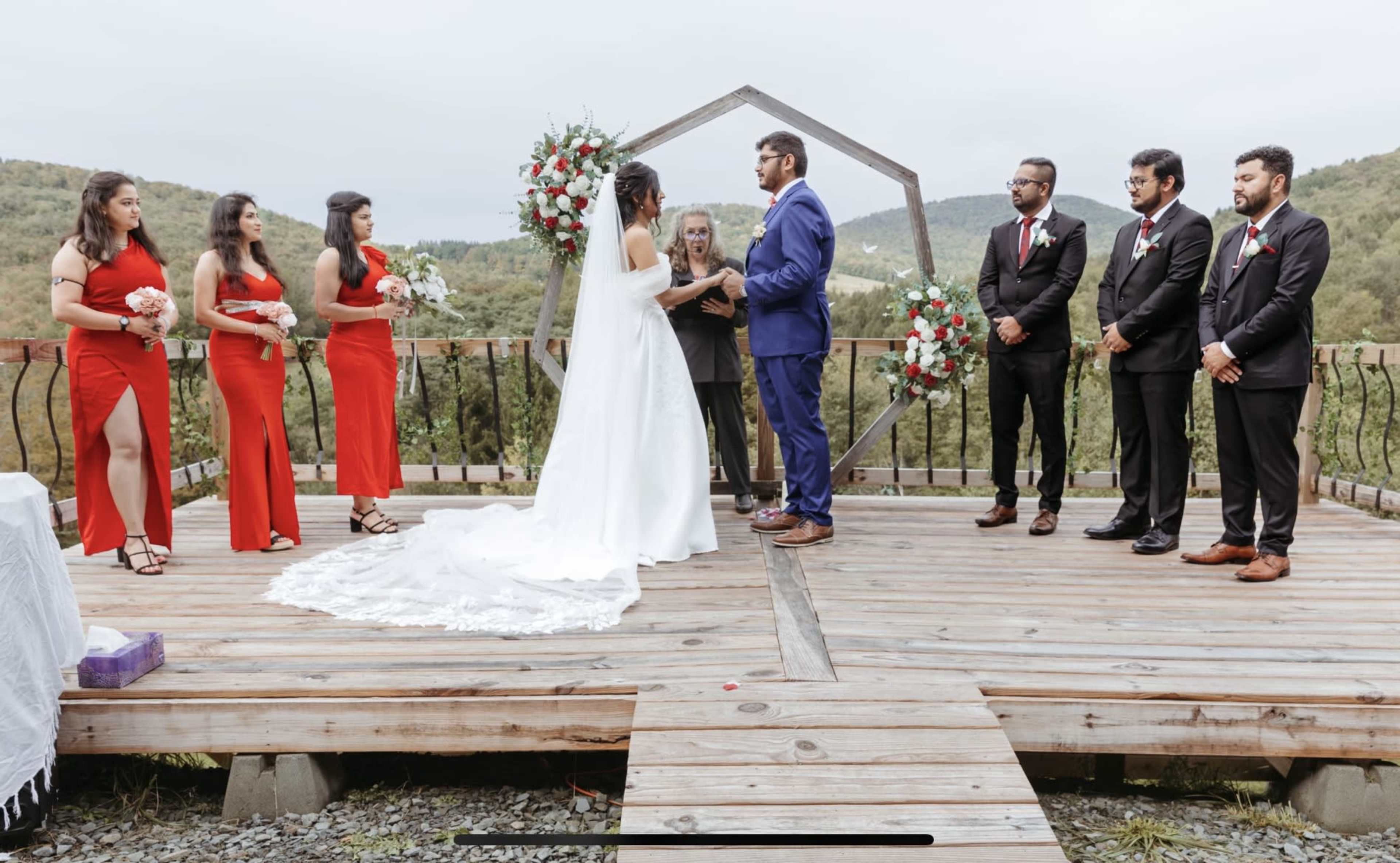 A couple exchanges vows under a decorative arch surrounded by their wedding party on a wooden platform in a scenic outdoor setting.