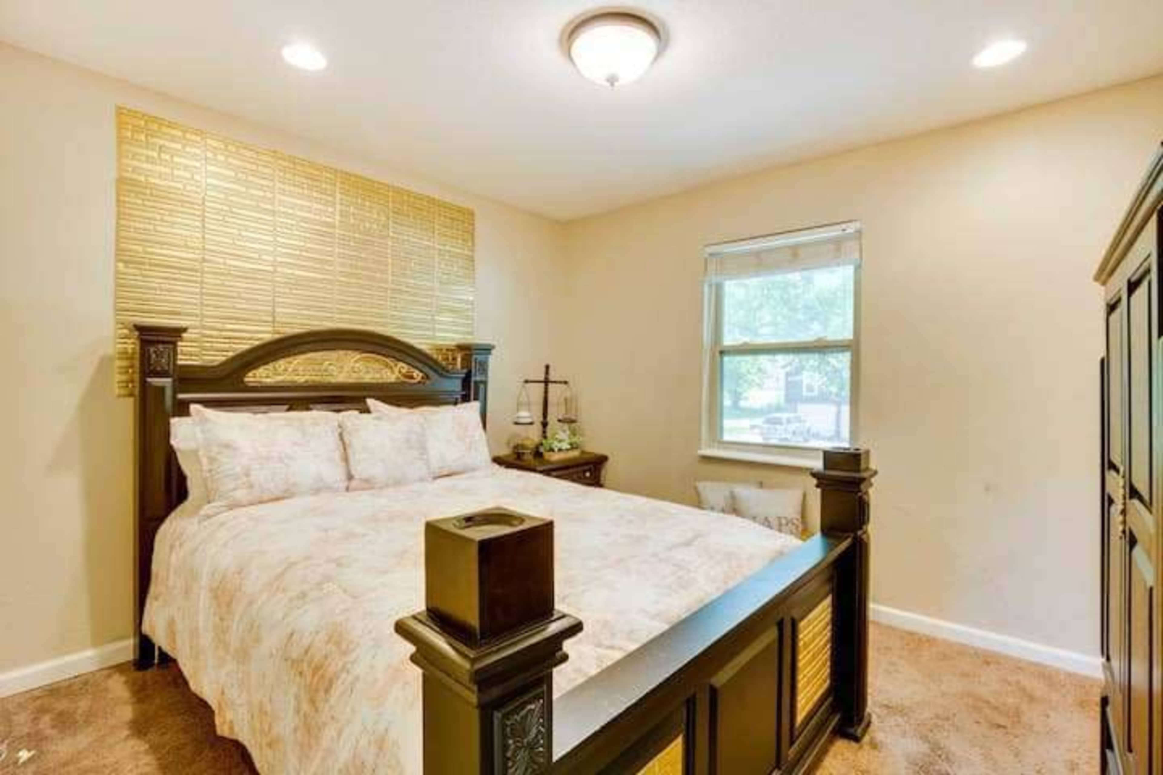 A cozy bedroom featuring a large bed with a decorative headboard, a window with blinds, and light-colored walls.
