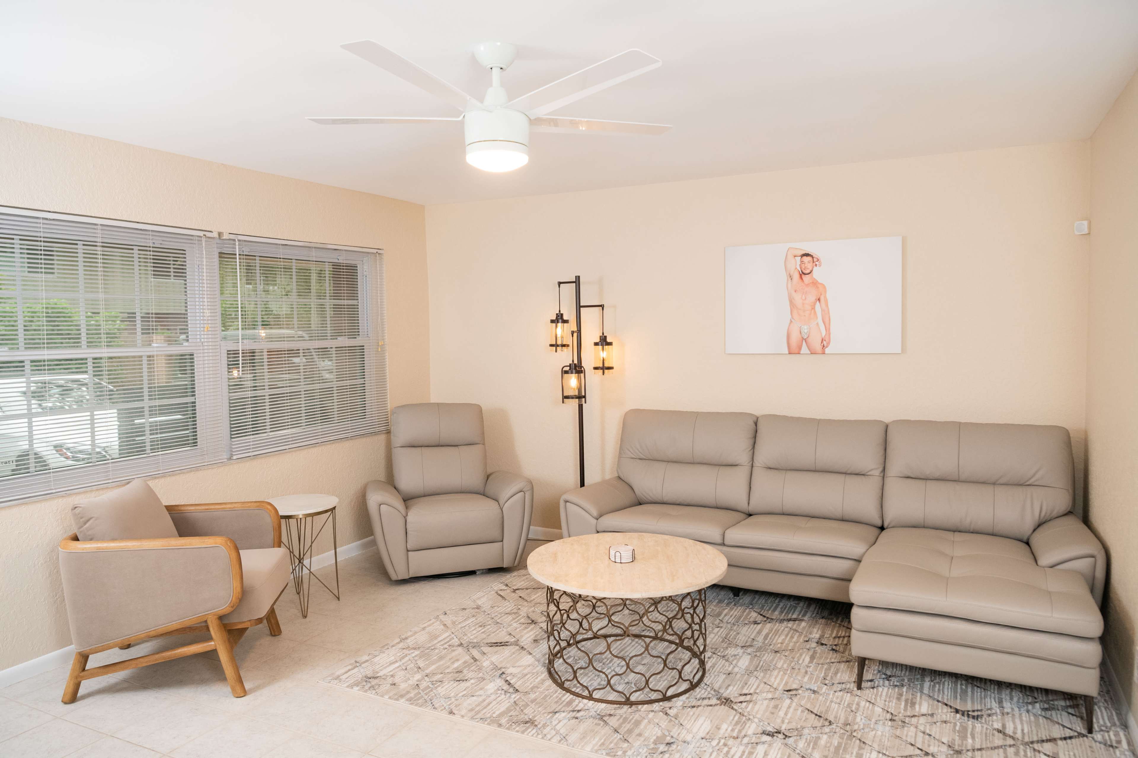The living room features a sectional sofa, an armchair, a round coffee table, and a wall art piece, along with large windows that allow natural light.