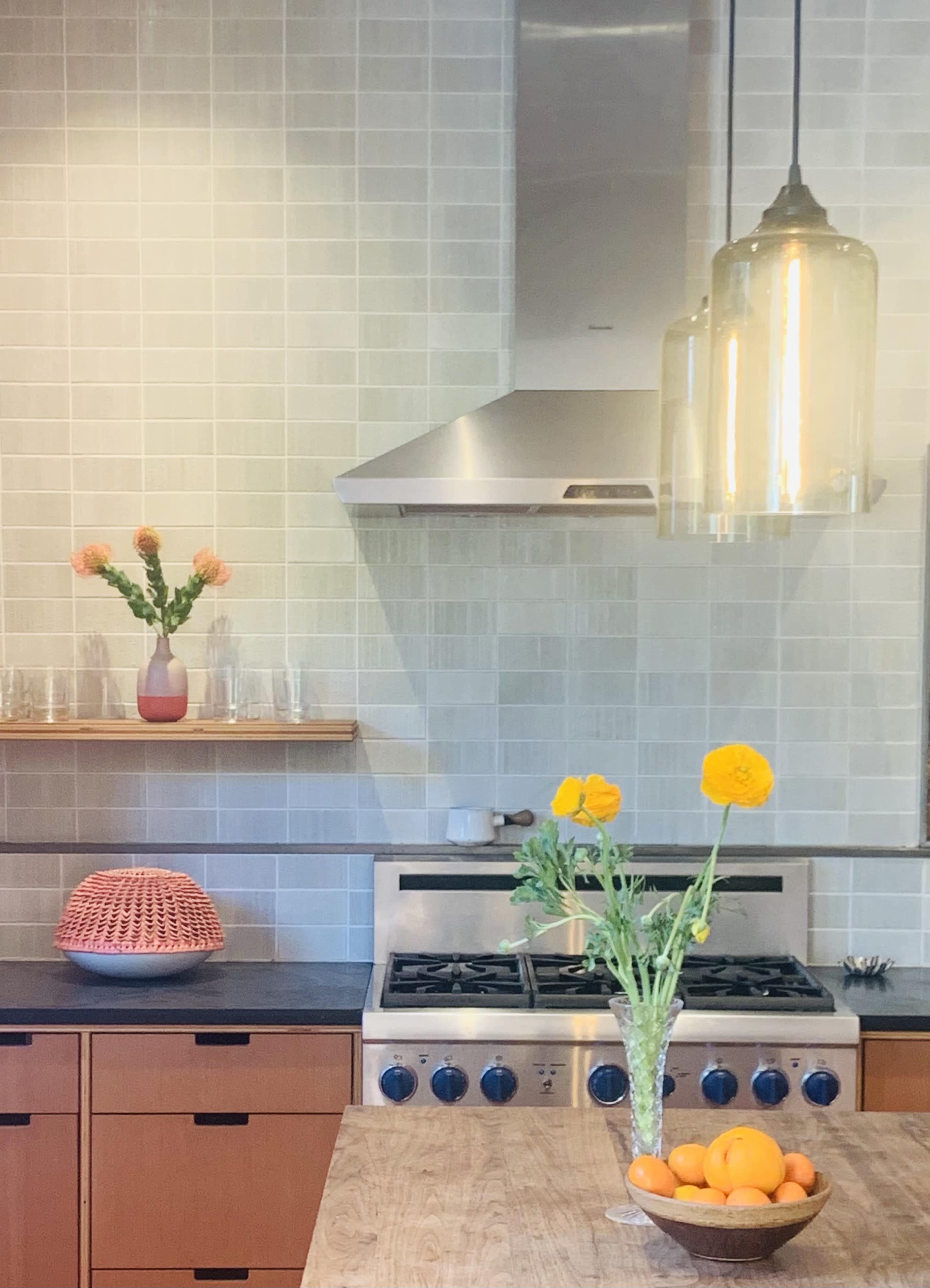 A modern kitchen features a stainless steel stove, a wooden table with a bowl of oranges, and a vase with flowers on the counter.