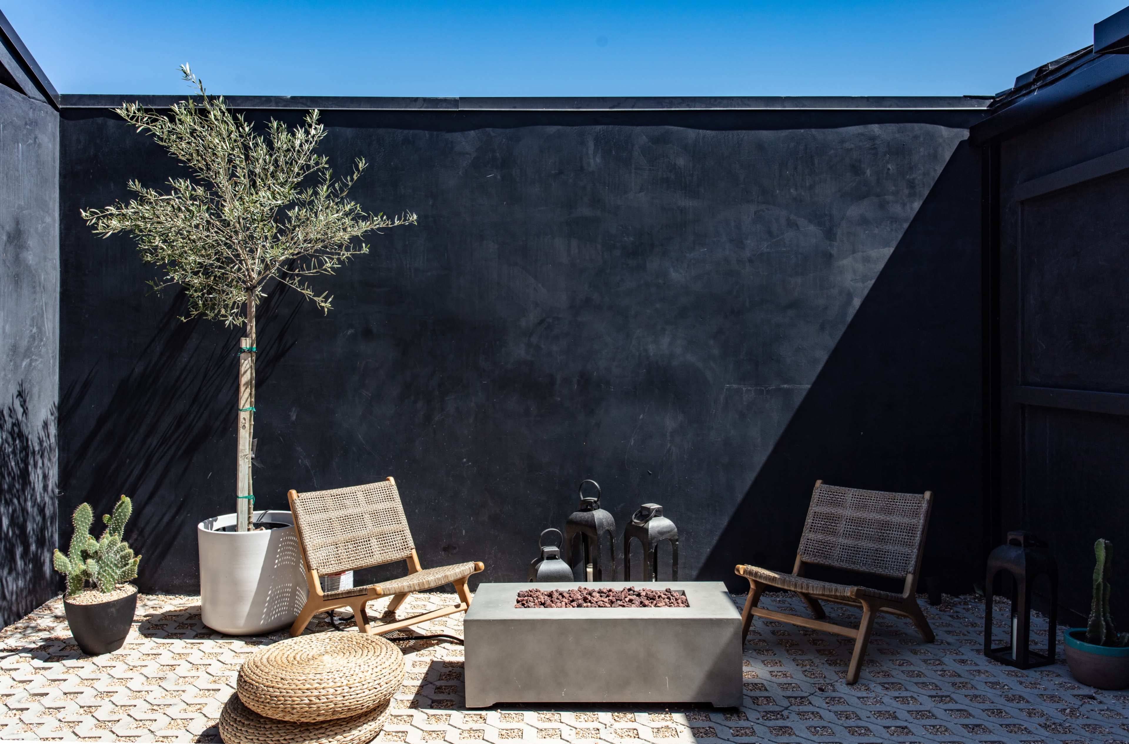 The scene features a minimalist outdoor patio with two wooden chairs, a low stone table surrounded by decorative items, a potted olive tree, and various plants against a dark wall.