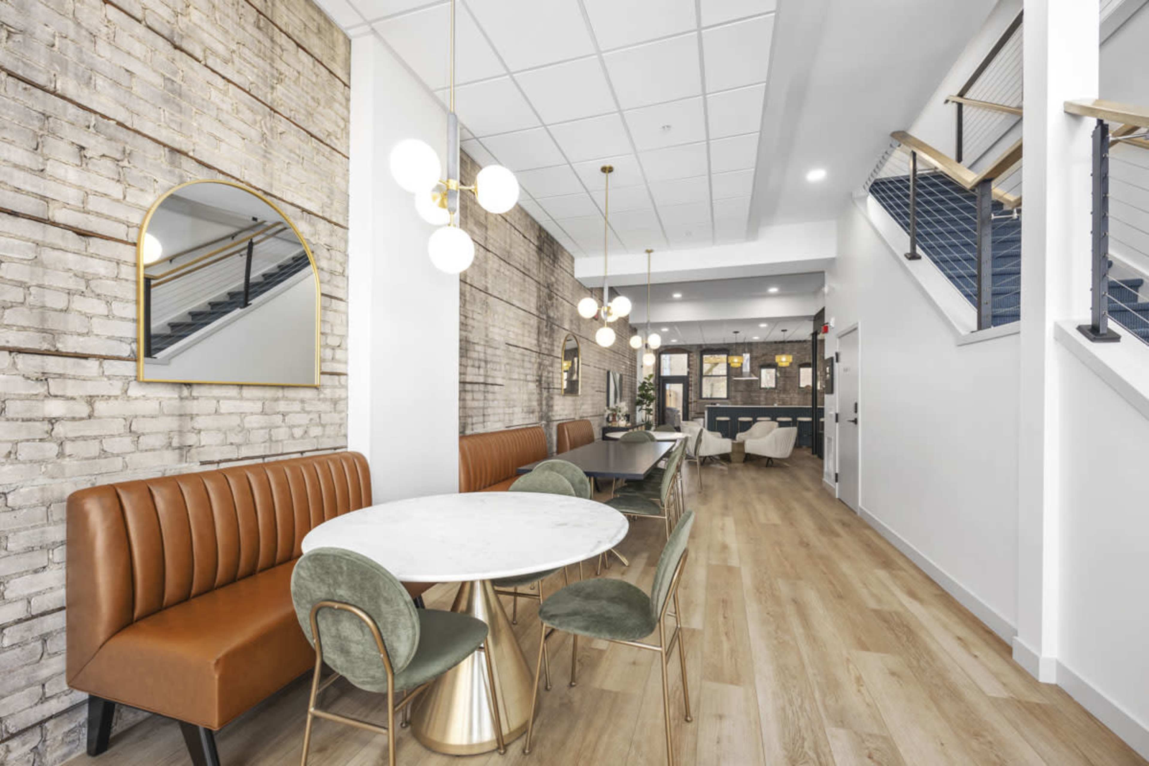 The image shows a modern interior space featuring a combination of a brick wall, a circular mirror, a marble dining table surrounded by green and gold chairs, and a staircase in the background.