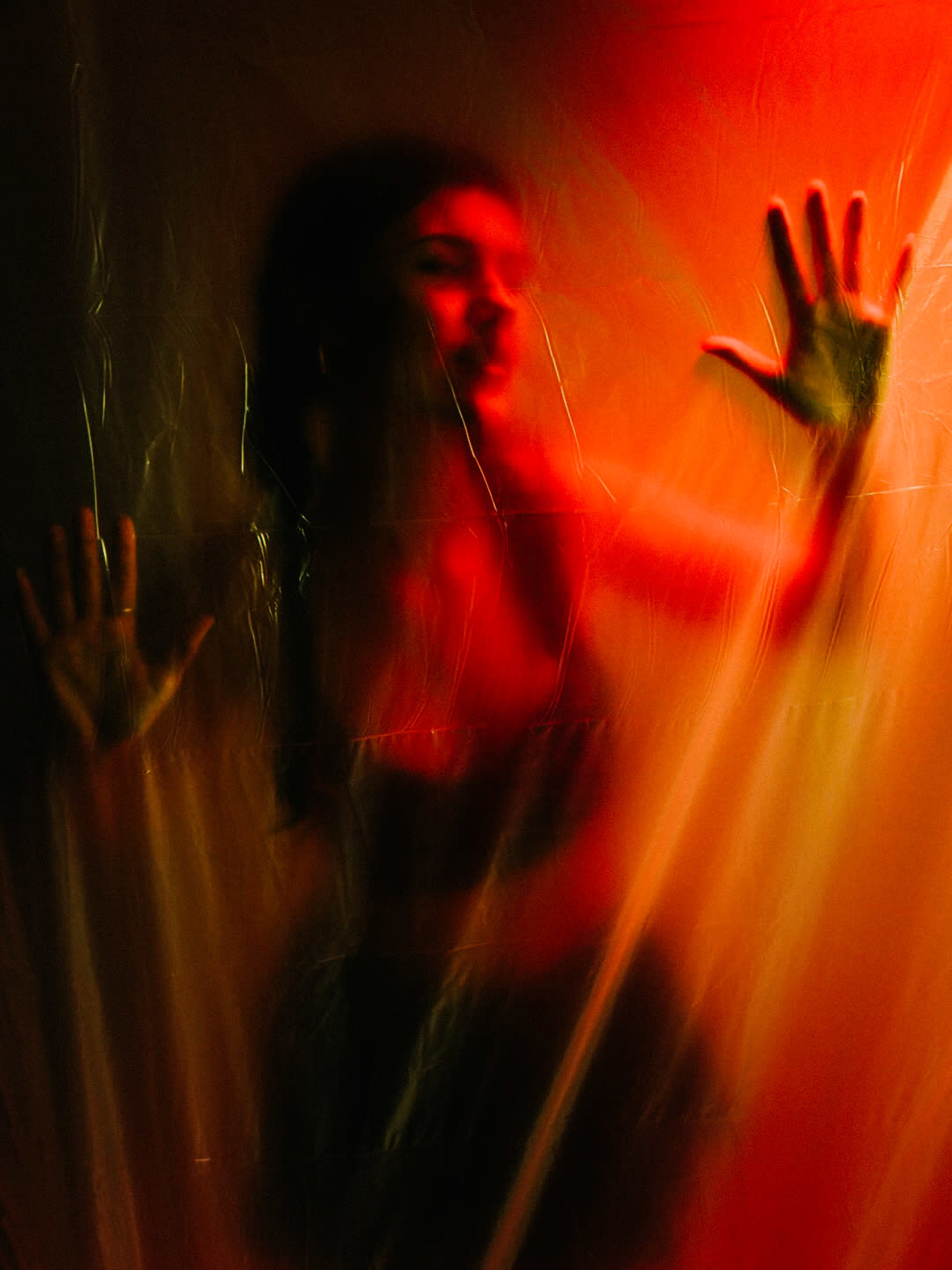 A figure is seen behind translucent plastic, illuminated by red and orange lighting, with hands pressed against the surface.