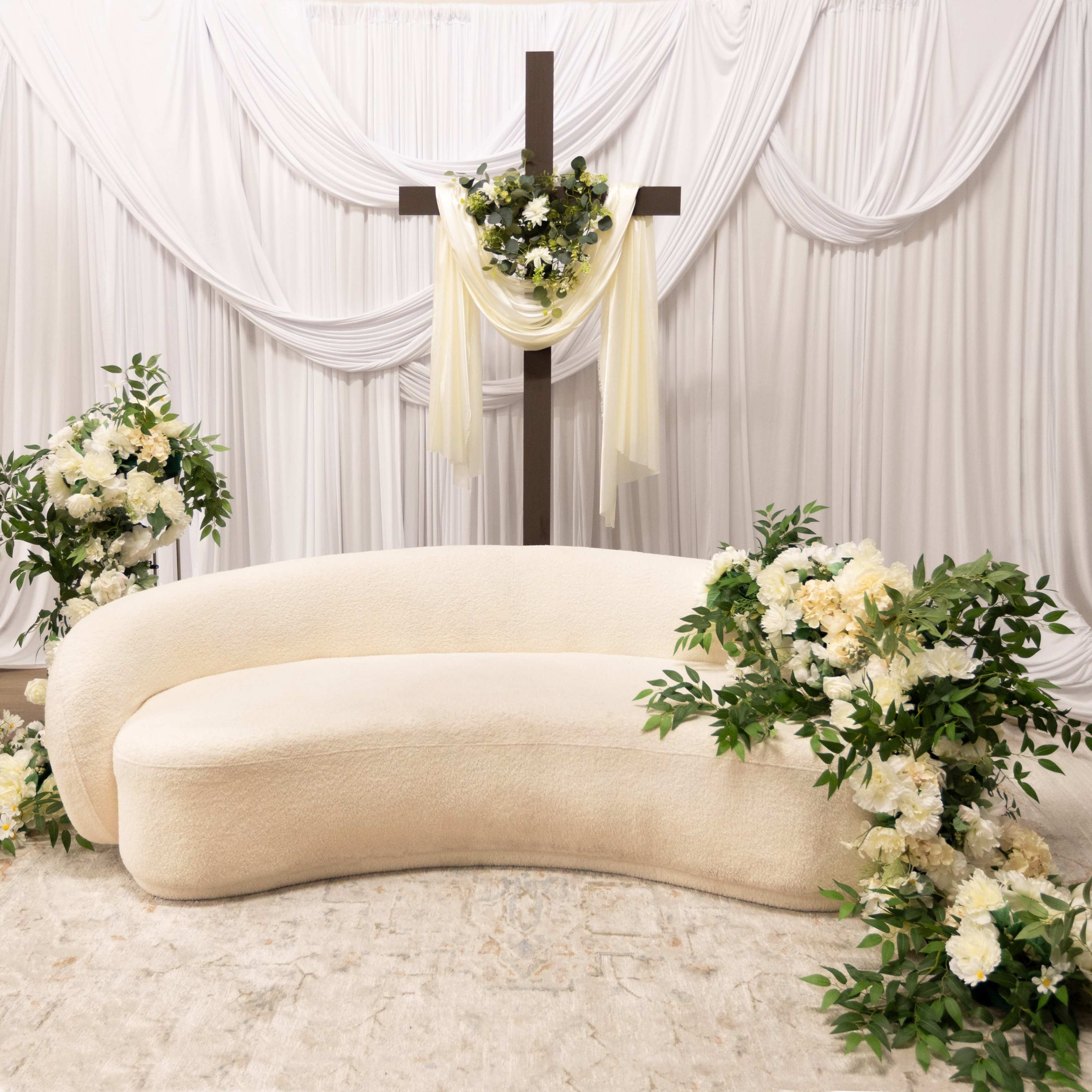 A curved cream-colored sofa is positioned in front of a decorative wooden cross adorned with flowers and draped fabric, surrounded by floral arrangements.