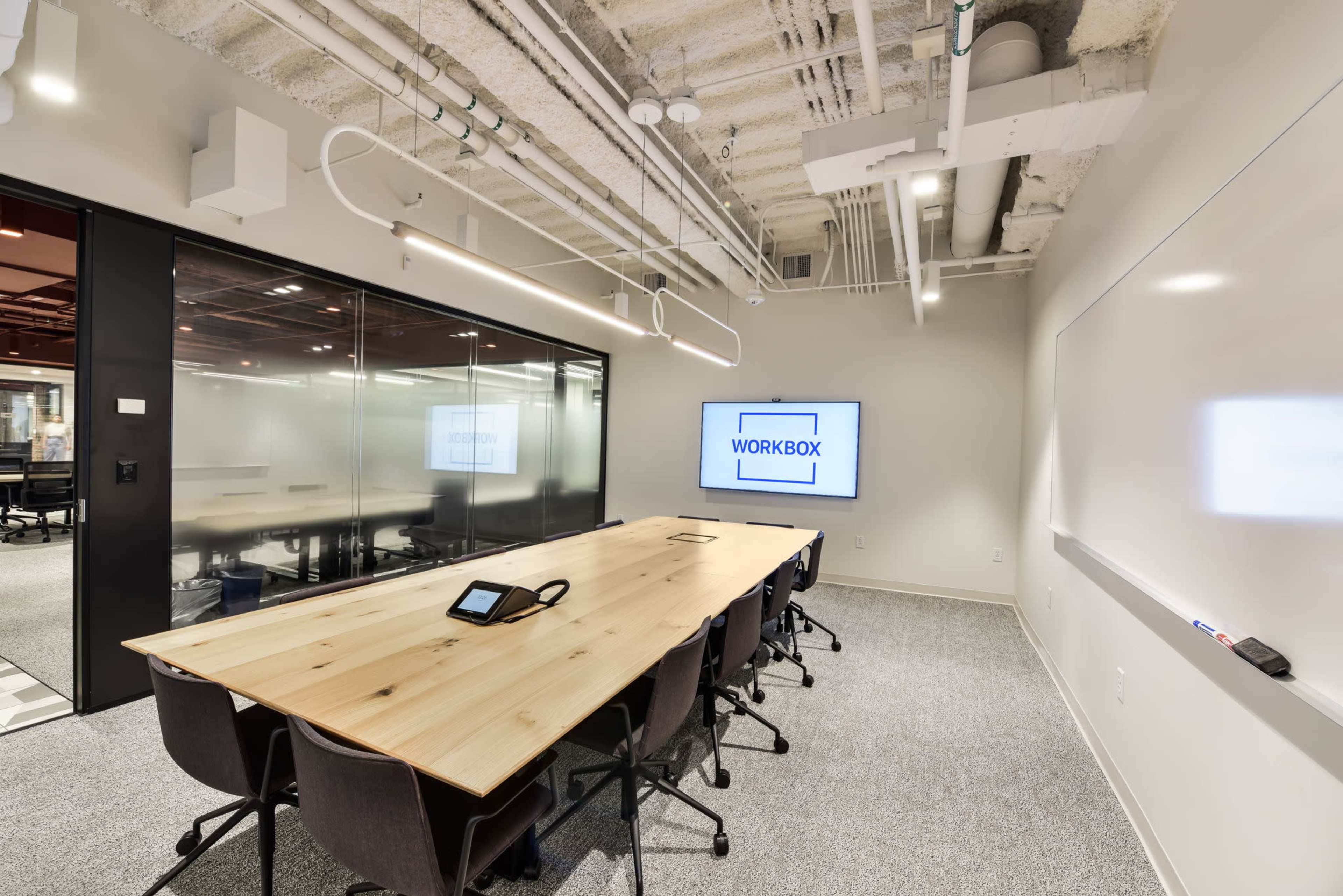 A modern conference room featuring a long wooden table, ergonomic chairs, a large display screen, and whiteboards on the walls.
