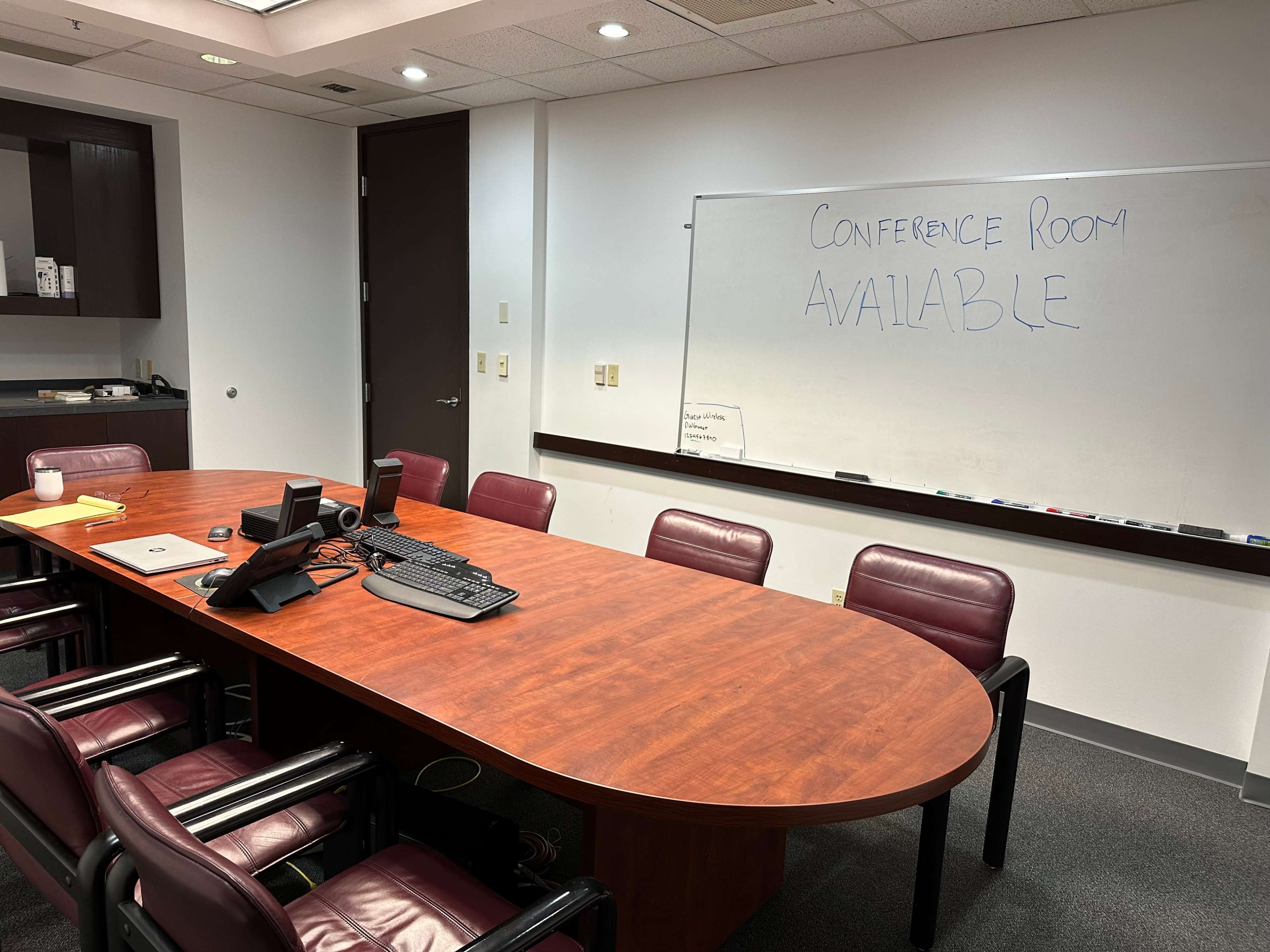 A large conference room features a wooden table surrounded by chairs, with a whiteboard displaying the message "CONFERENCE ROOM AVAILABLE."