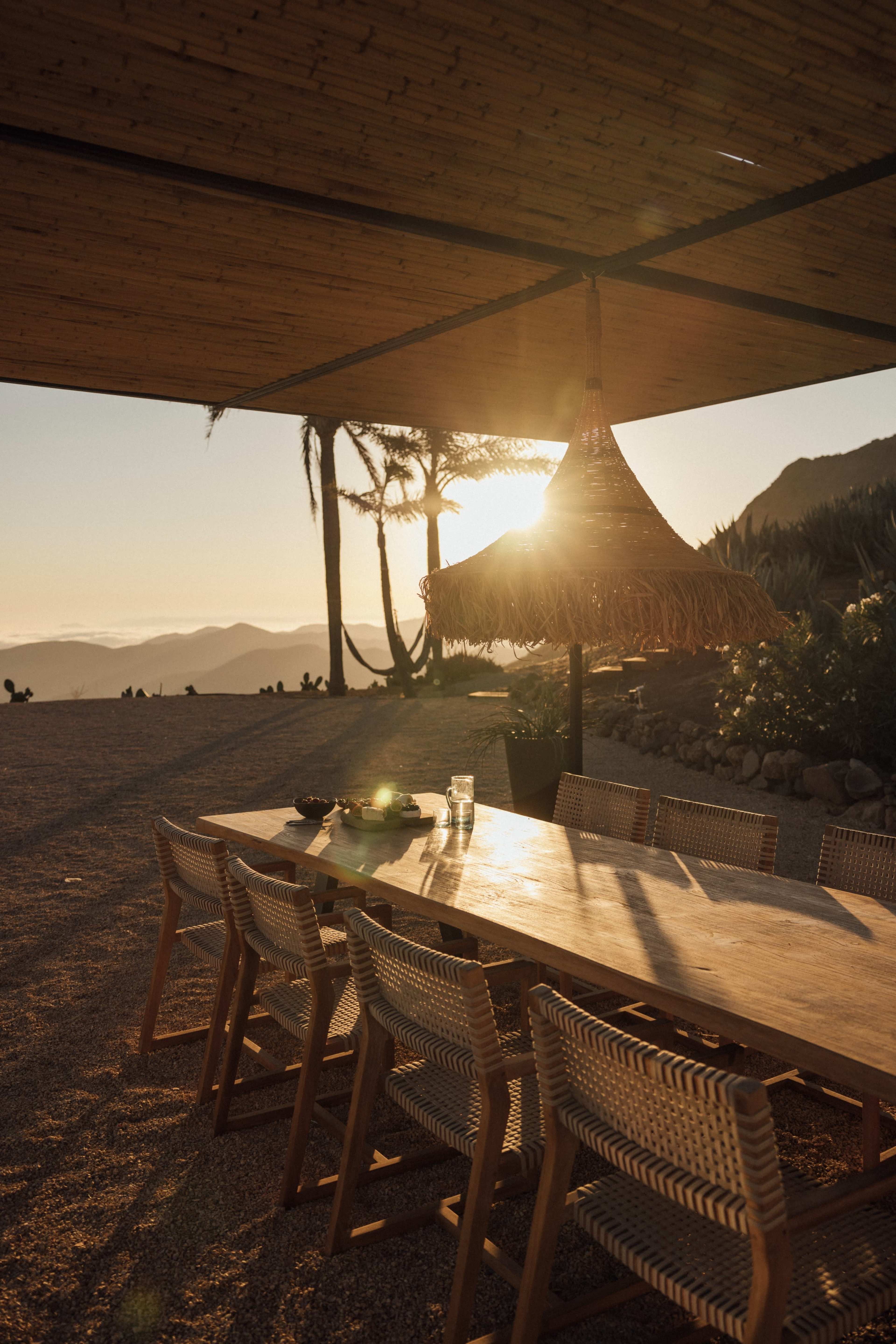 A large wooden table with woven chairs is set under a shaded area, with the sun setting in the background, illuminating the scene.
