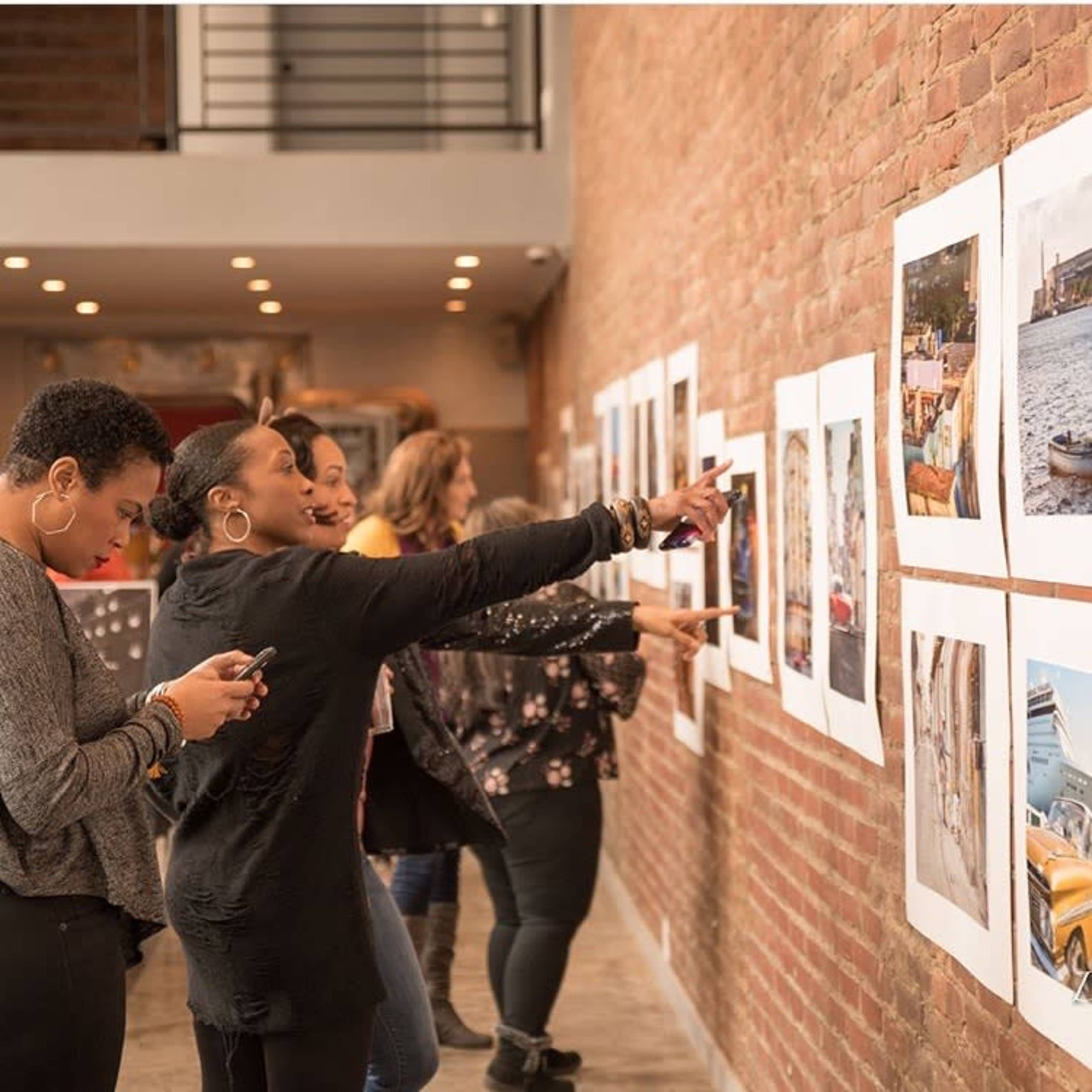 A group of women examines and discusses framed photographs displayed on a brick wall in an indoor gallery.