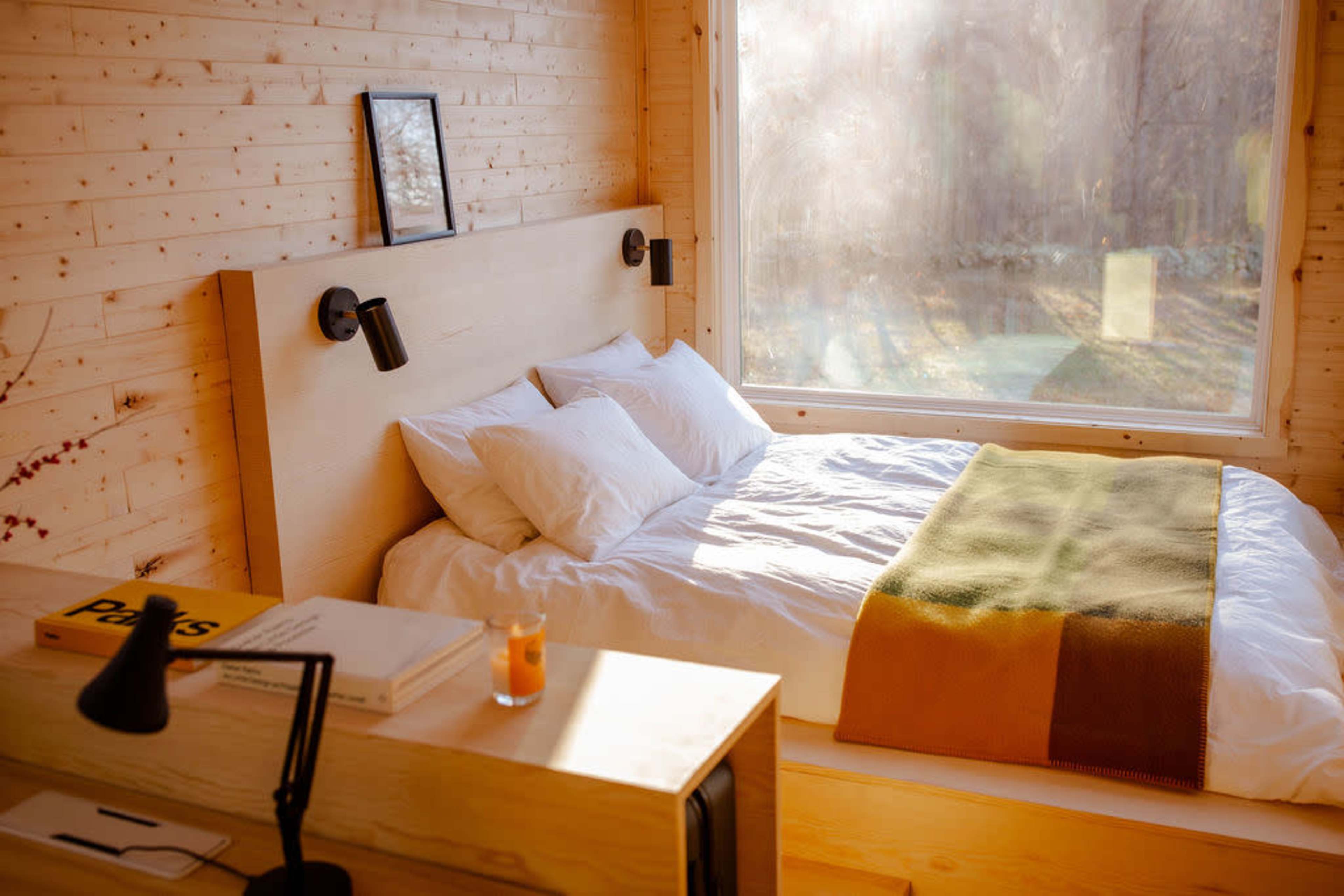 A cozy bedroom with a wooden wall, a large window allowing natural light, and a neatly made bed with white linens and a green throw.