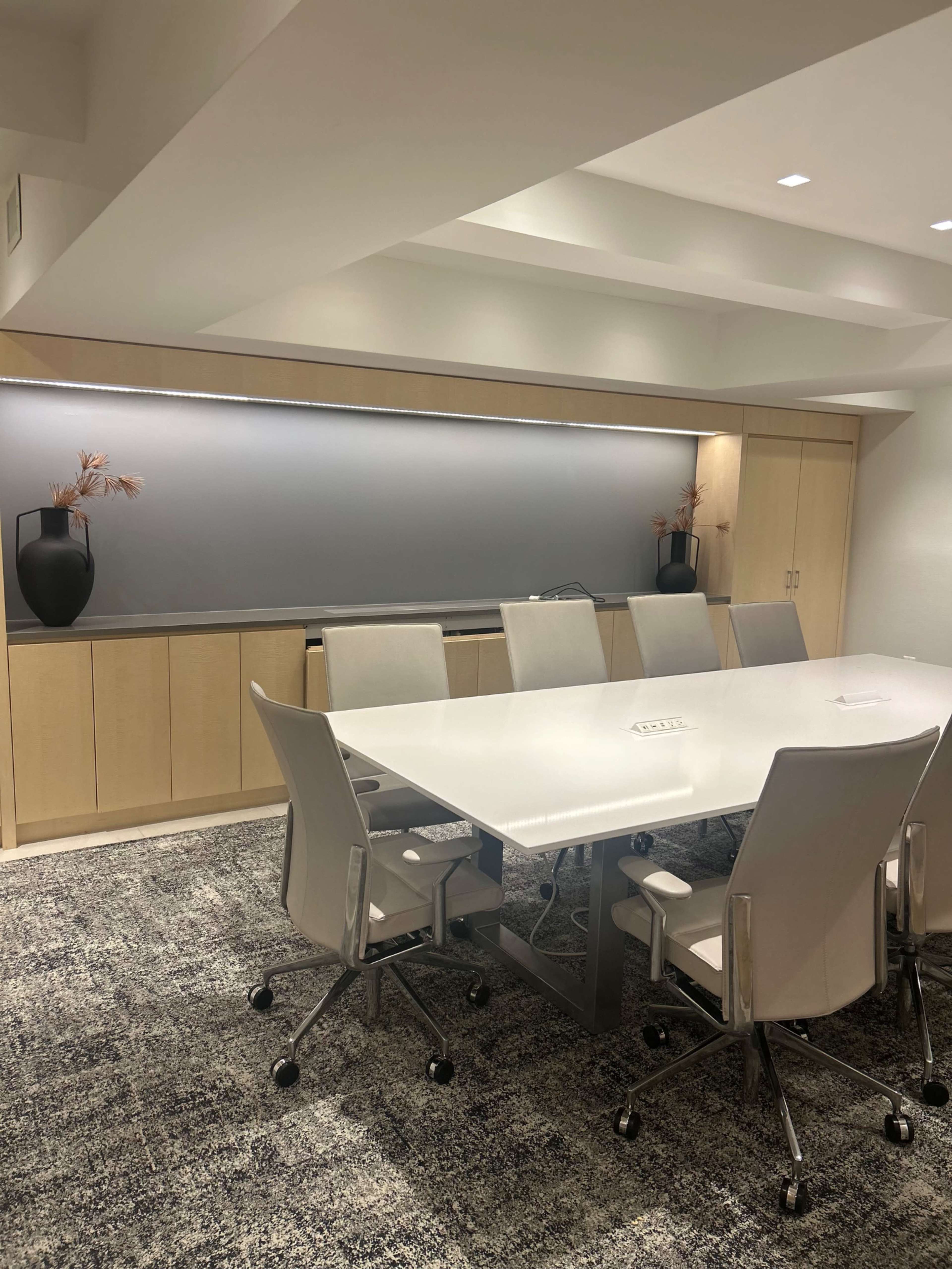 A sleek, modern conference room features a large white table surrounded by eight light-colored chairs, with a minimalist cabinet and decorative vases in the background.
